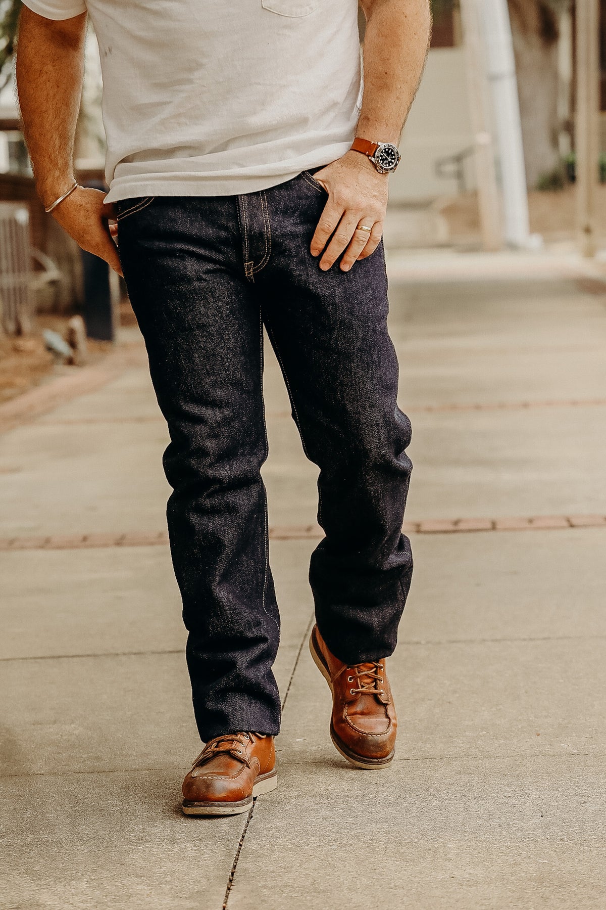 A person standing on a sidewalk wearing a white t-shirt and the 21 oz Selvedge Denim Super Slim Tapered Jeans in indigo color with brown shoes.