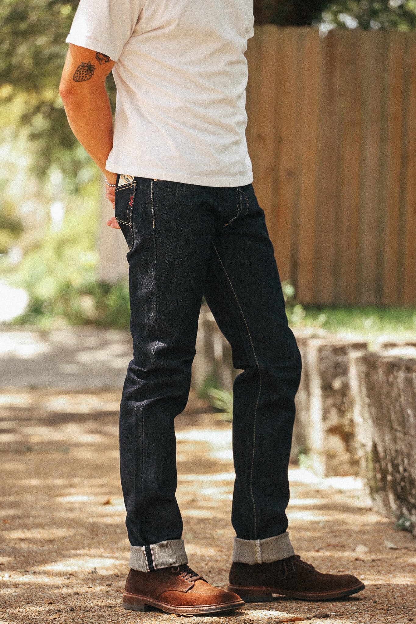A person wearing dark indigo denim jeans with rolled cuffs, paired with a white t-shirt and brown leather shoes, standing outdoors on a paved path with a wooden fence in the background.