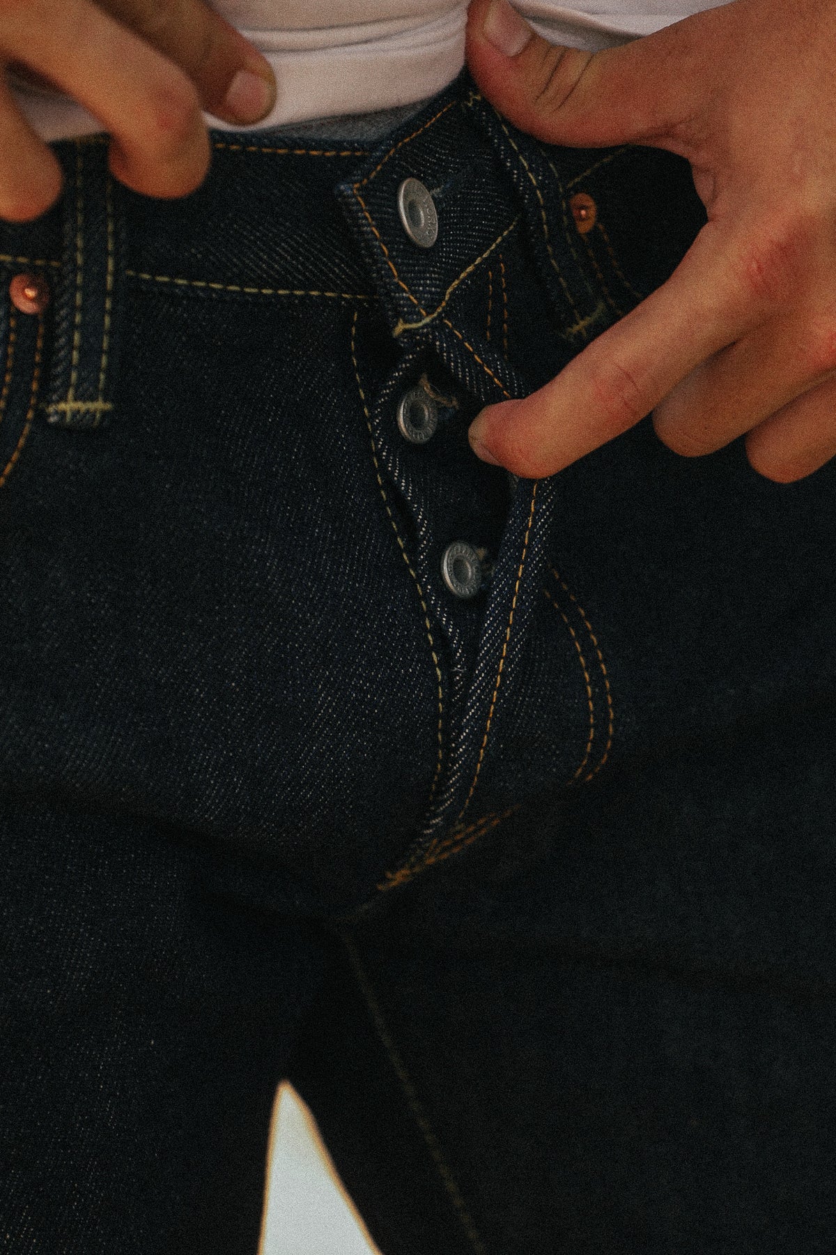 Close-up of a person wearing dark indigo denim jeans, pulling the waistband slightly outward with both hands to show the fit and button closure.