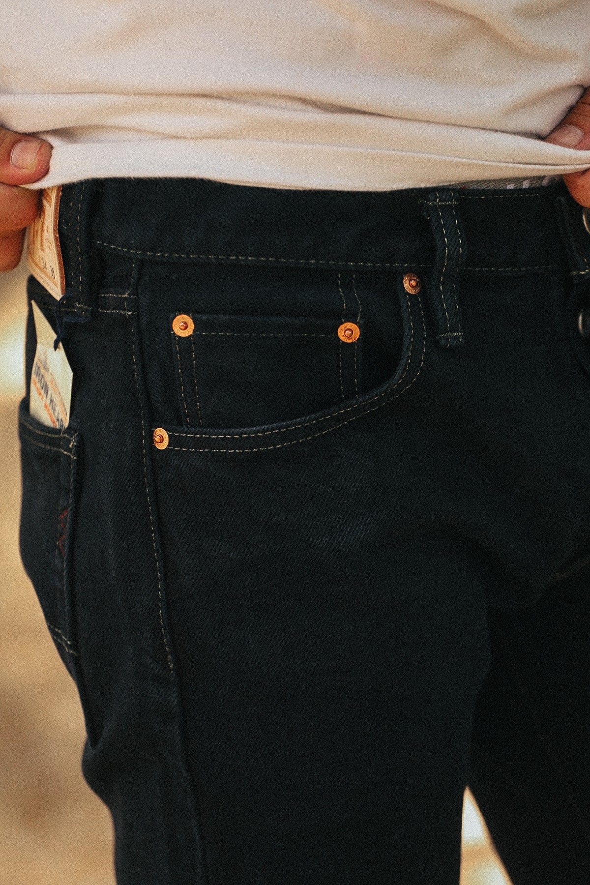 Close-up of a person wearing dark indigo denim jeans with copper-toned rivets and a cream-colored t-shirt being pulled up.