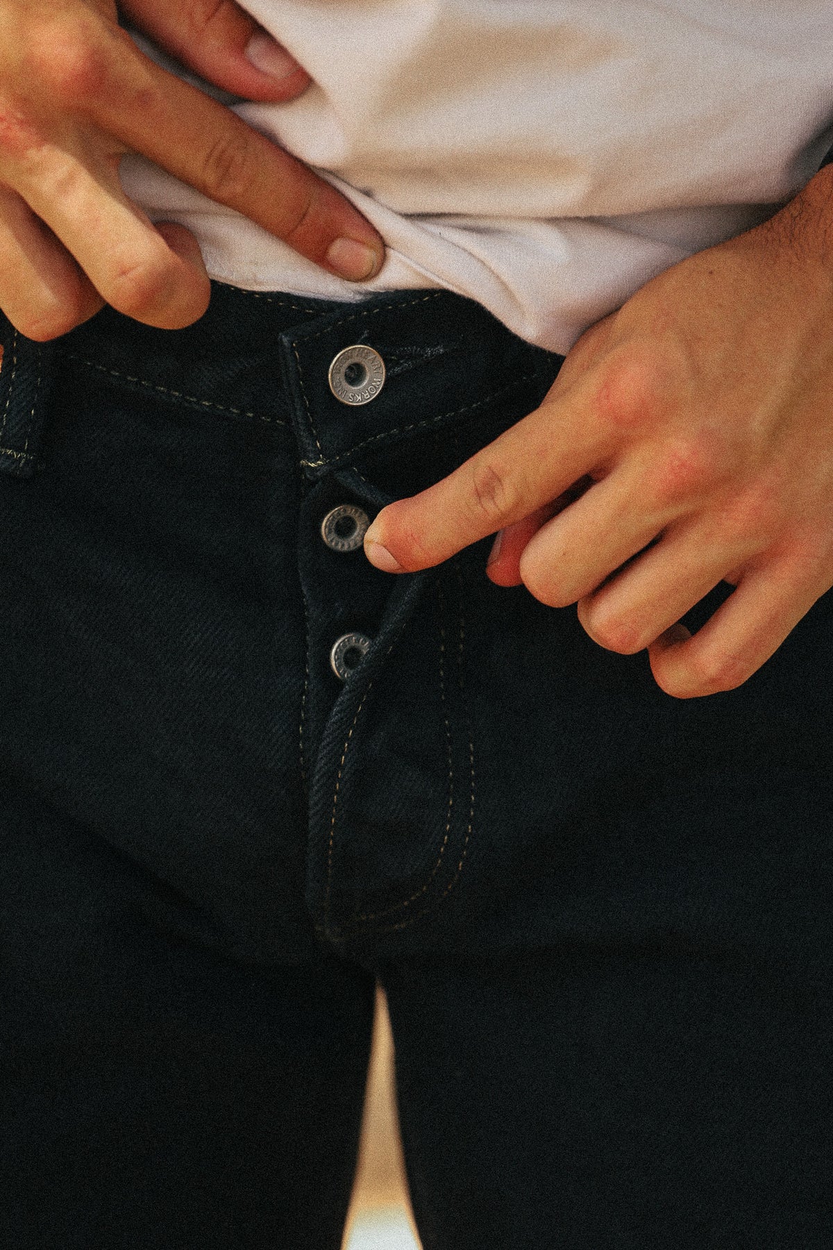 Close-up of a person wearing dark indigo jeans with a white t-shirt, showing the button closure of the jeans being adjusted by hands.