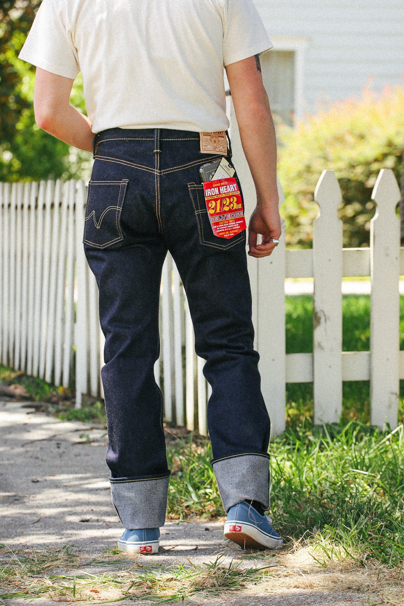 A person viewed from behind wearing dark blue denim jeans with cuffed hems, a cream t-shirt, and blue sneakers, standing outdoors on a path next to a white picket fence.