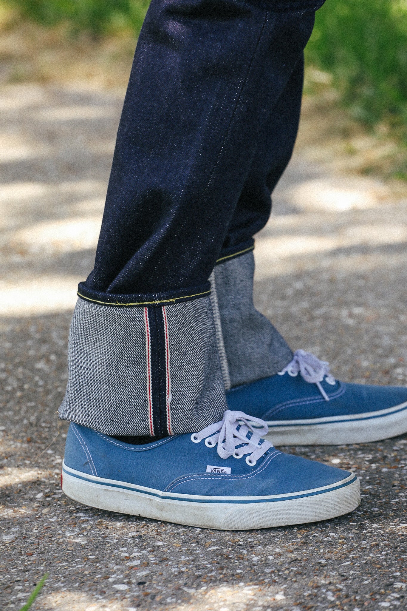 A close-up of a person's legs wearing dark indigo denim jeans with the cuffs turned up to show the raw, textured weave, paired with blue canvas low-top sneakers on an outdoor gravel path.