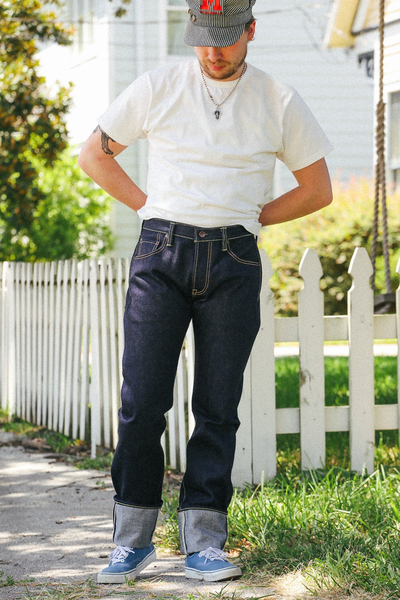 A man wearing a white t-shirt, dark blue cuffed jeans, a gray baseball cap, and blue sneakers, standing outdoors in front of a white picket fence.