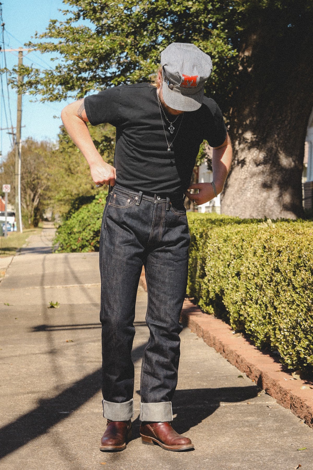A man wearing a black t-shirt, dark wash jeans with rolled cuffs, brown leather boots, and a gray cap with a red logo, standing on a sunny sidewalk next to a tree.