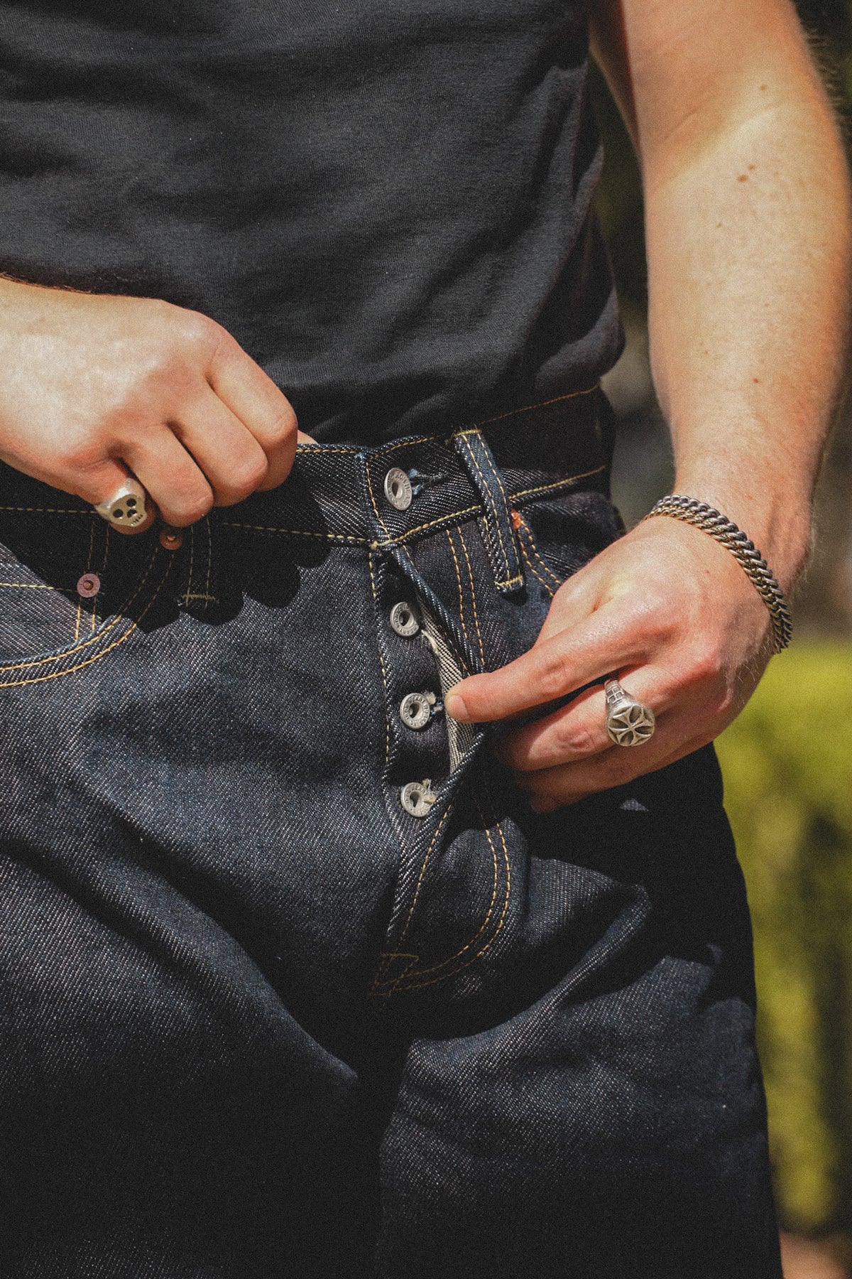 Close-up of a person wearing dark indigo denim jeans with a button fly, pulling the fly open with both hands. The person is also wearing a black t-shirt, silver chain bracelet, and silver rings.
