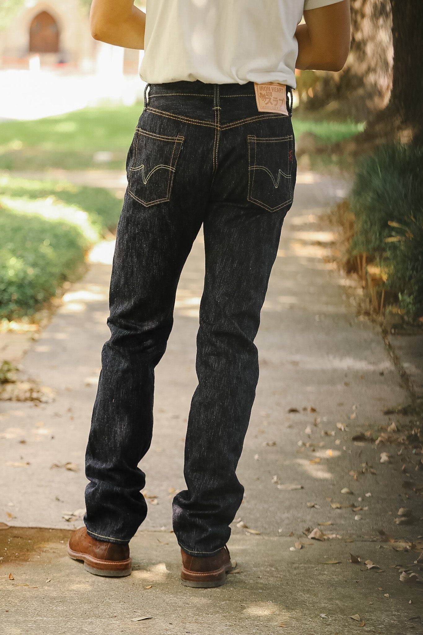 Rear view of a person wearing dark indigo denim jeans and brown boots on an outdoor paved path.