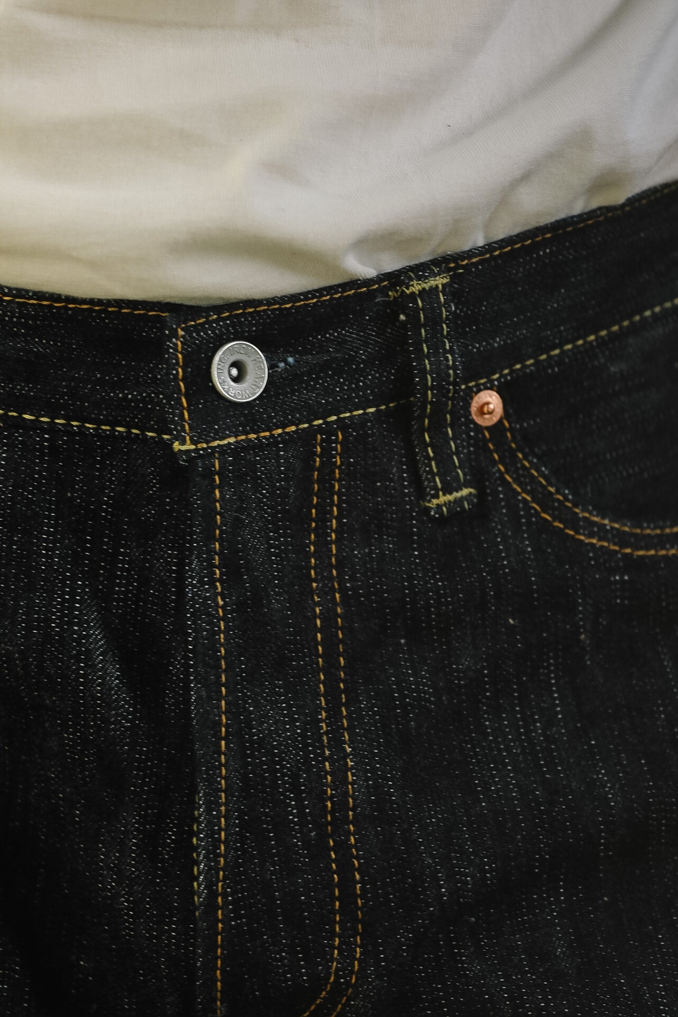 Close-up of dark indigo denim jeans with yellow contrast stitching, a silver metal button, and a copper-toned rivet, worn with a light-colored shirt.