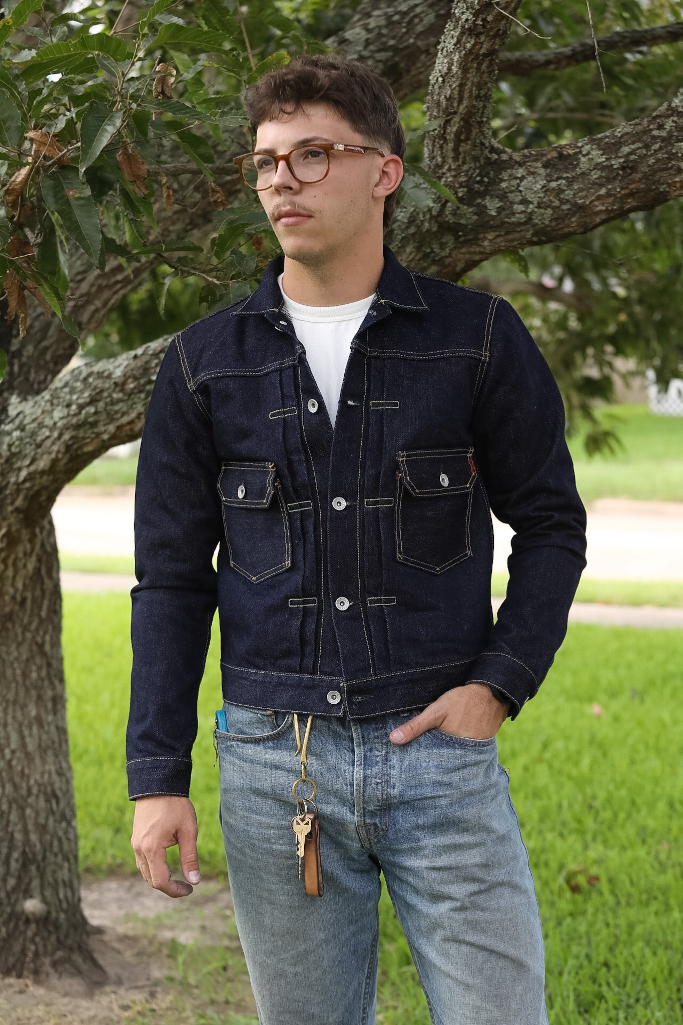 A man wearing a dark indigo denim jacket over a white t-shirt and light blue jeans, standing outdoors in front of a tree.
