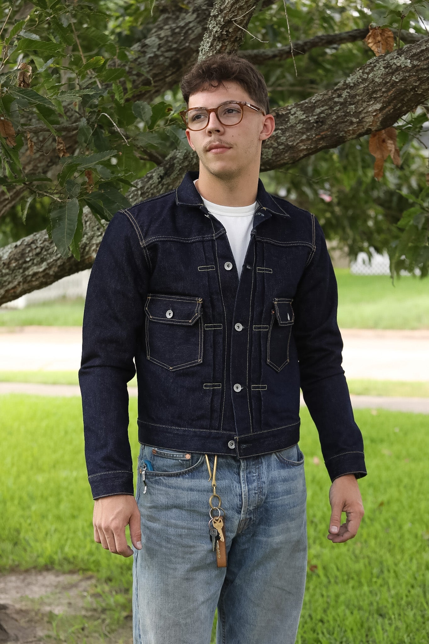 A man wearing a dark indigo denim jacket over a white t-shirt and light blue jeans, standing outdoors in front of a tree with green grass in the background.