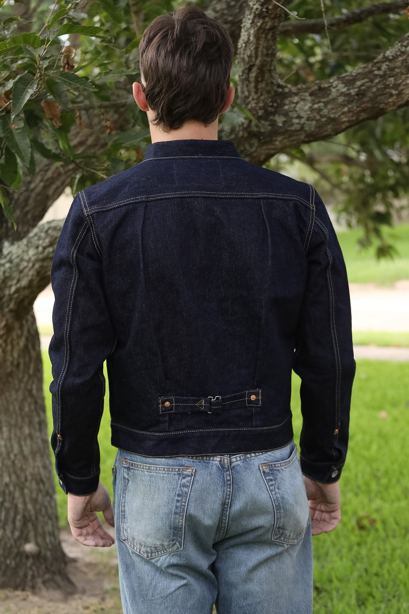 A man viewed from behind wearing a dark navy blue denim jacket with contrast stitching and light blue jeans, standing outdoors in front of a tree.