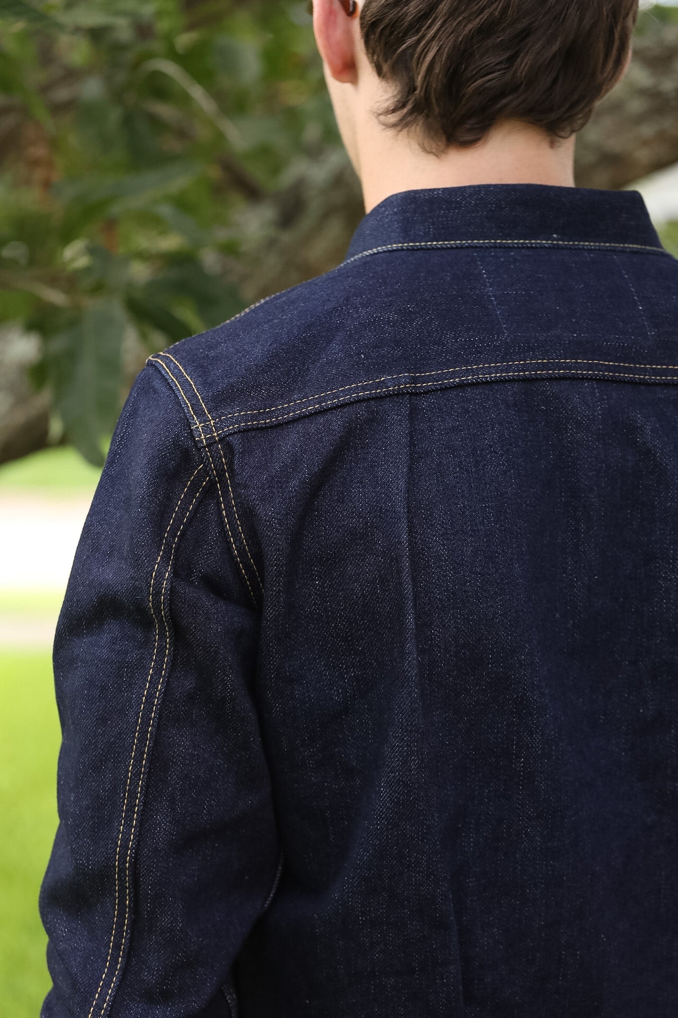 Back view of a person wearing a dark indigo denim jacket outdoors, showing the collar, shoulder seam, and contrast stitching against a blurred green background.