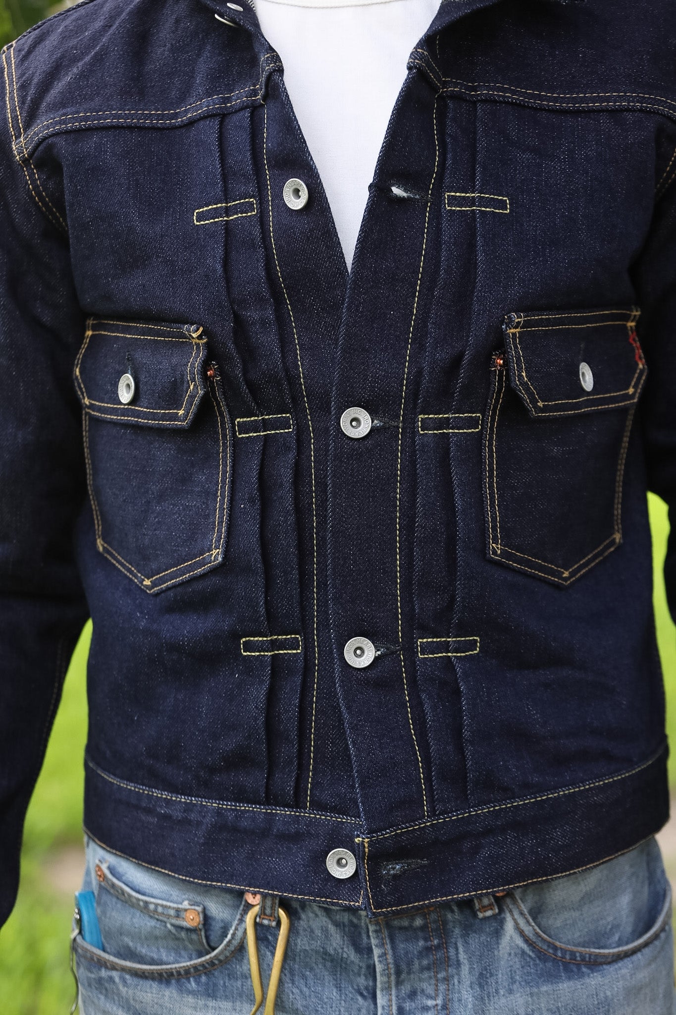 A close-up of a dark indigo denim jacket with contrast light stitching, silver snap buttons, and two front flap pockets, worn over a white shirt and blue jeans.