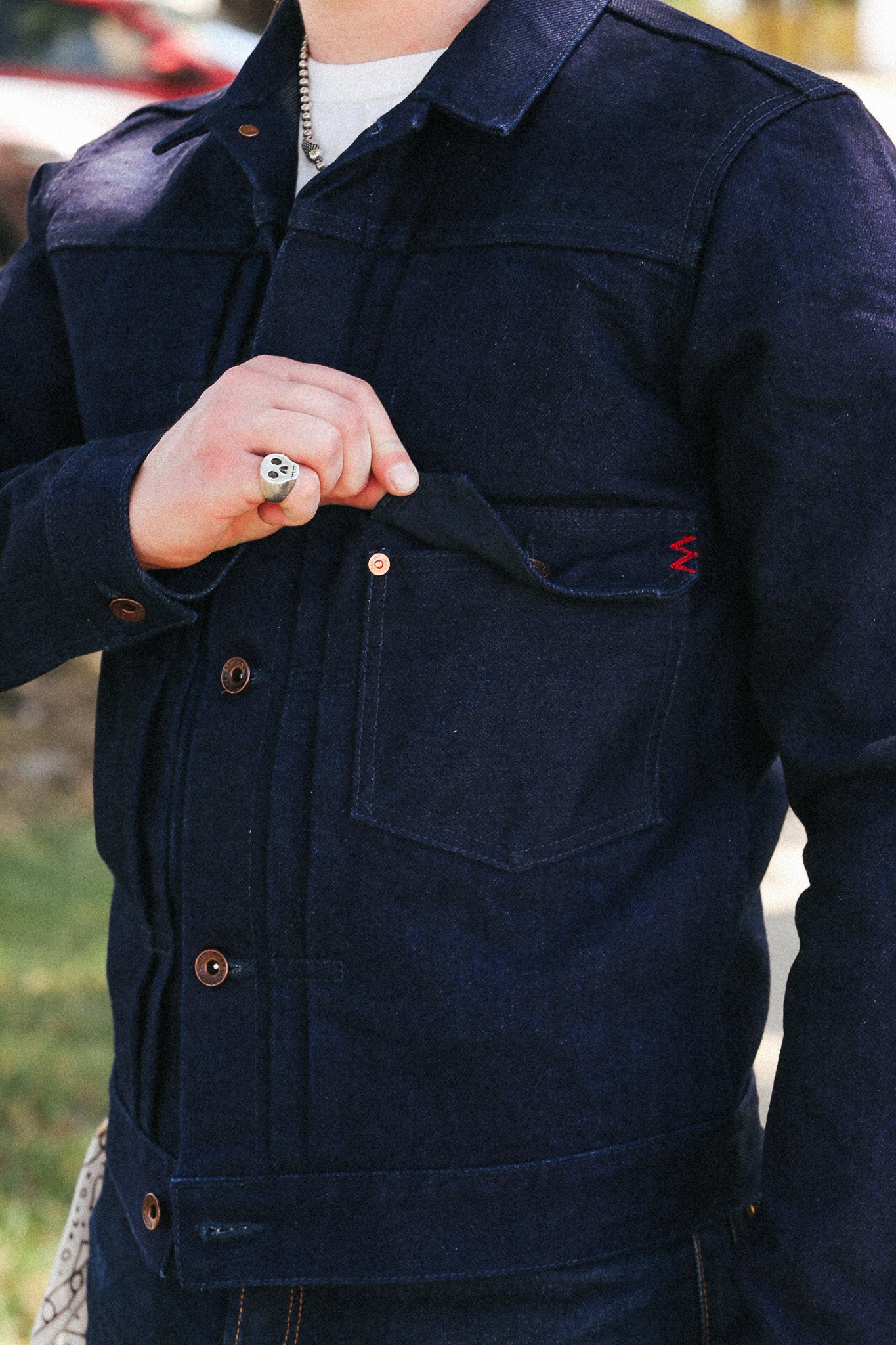 A close-up of a person wearing a dark indigo denim jacket, showing the chest pocket with a red zigzag embroidery detail and brown buttons.