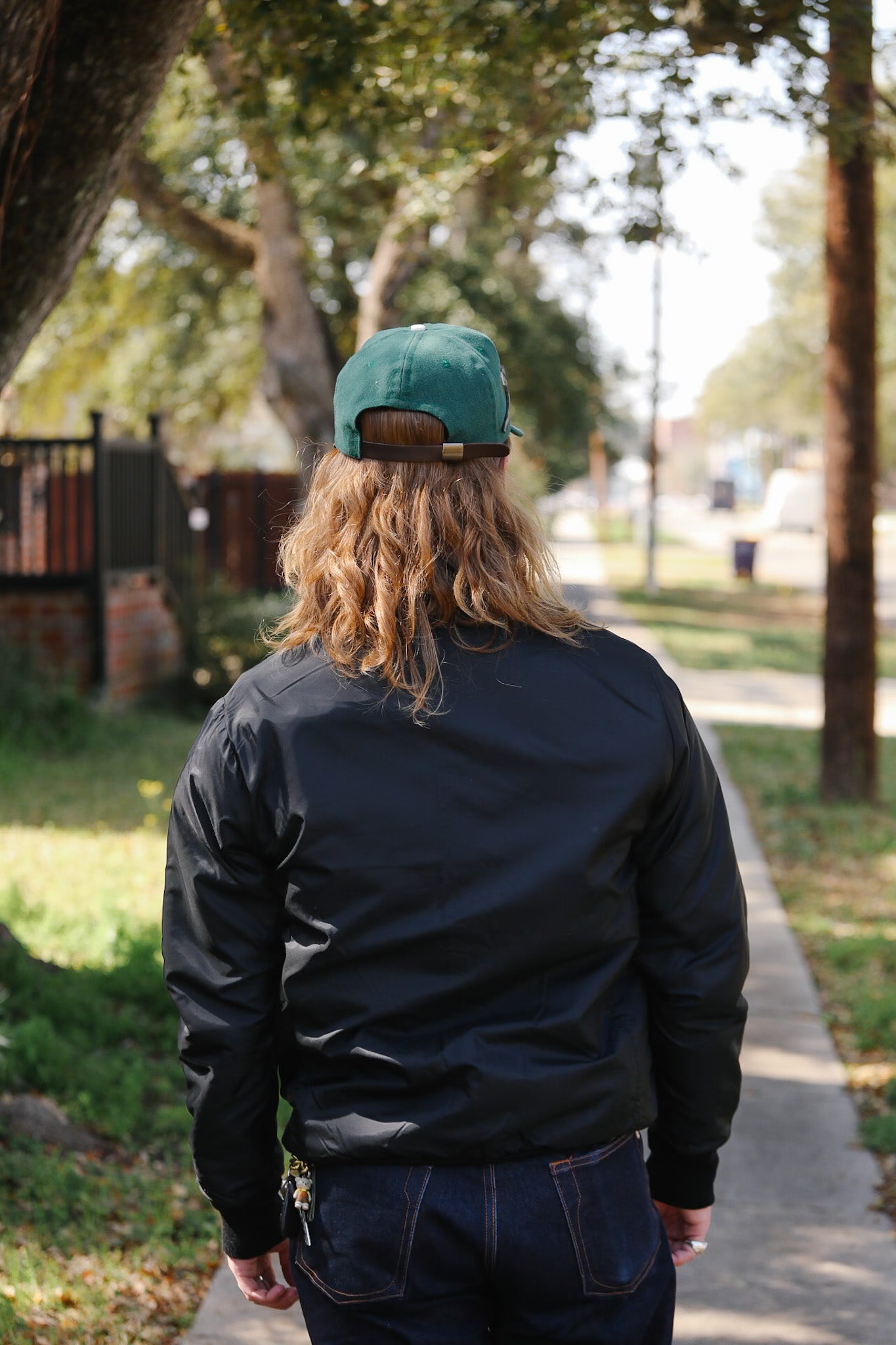 Person wearing a teal cap and dark jacket walking away on a path with trees and grass in the background