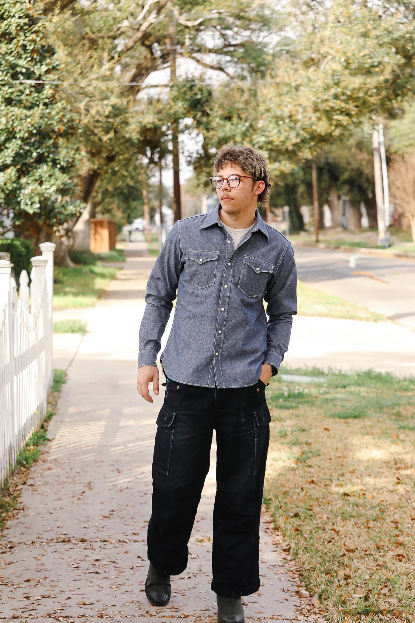 Man wearing a gray shirt and black pants walking on a sidewalk with trees and a house in the background.