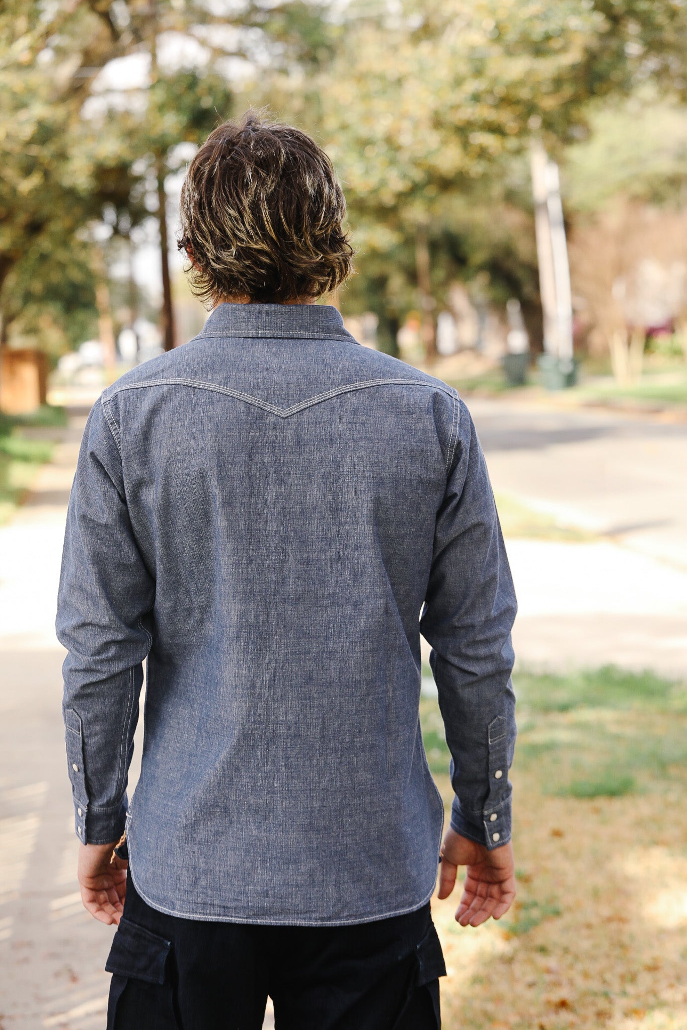 Person wearing a blue denim shirt walking outdoors with trees in the background