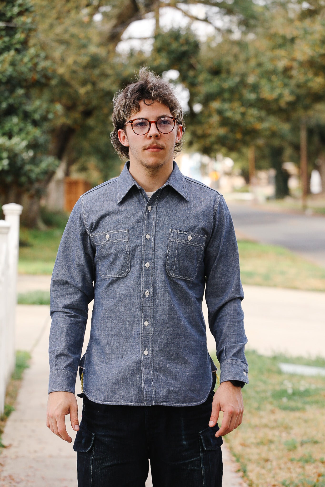 Person wearing a blue shirt standing outdoors with trees and a road in the background