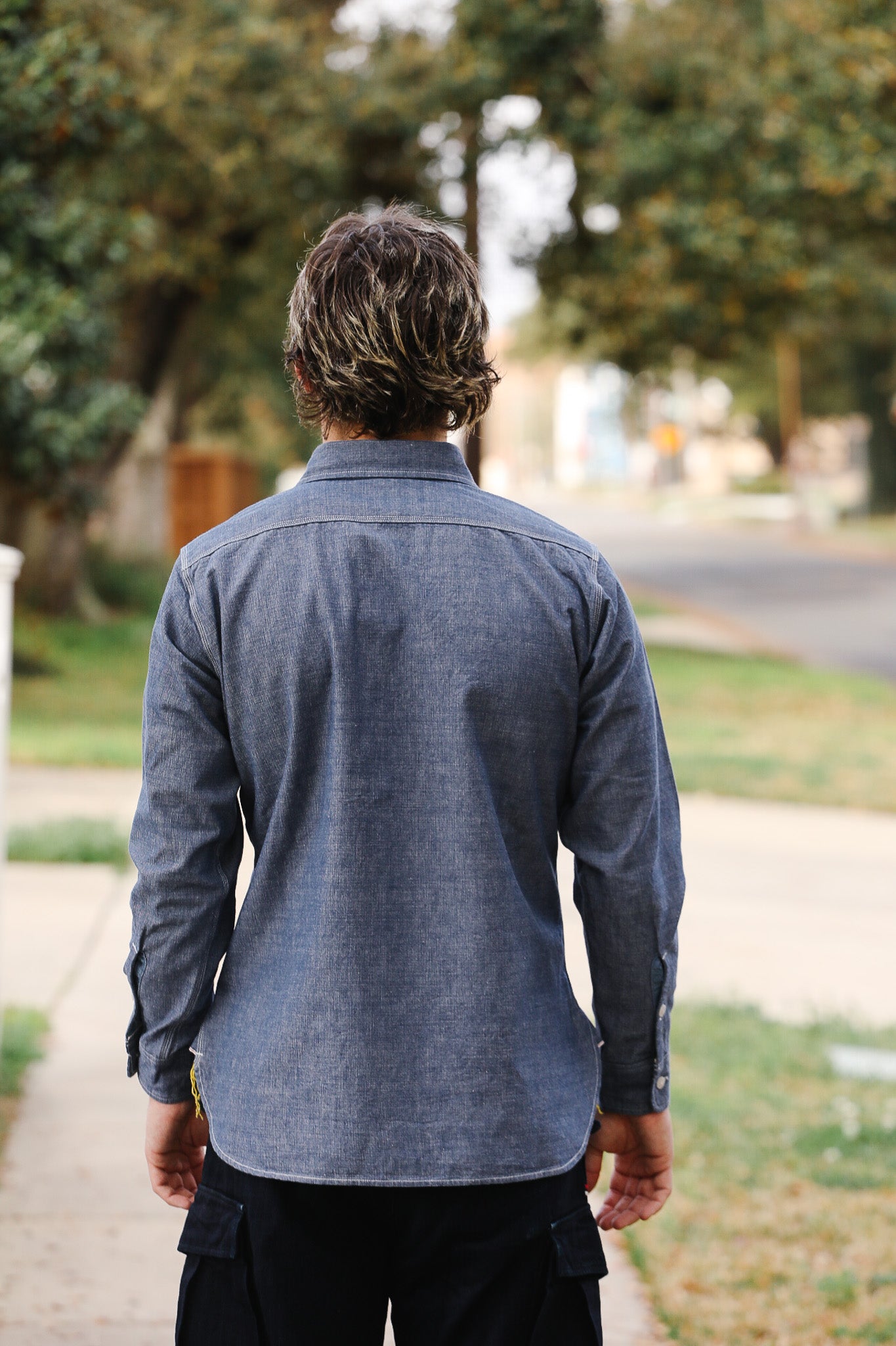 Person wearing a blue shirt walking on a sidewalk with trees and a road in the background