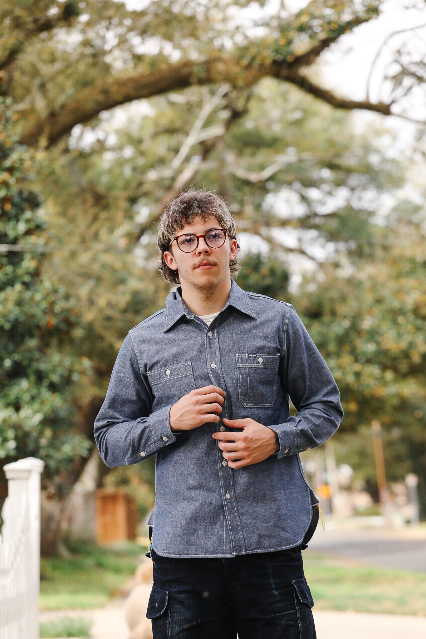 Person wearing a blue denim shirt standing outdoors with trees in the background