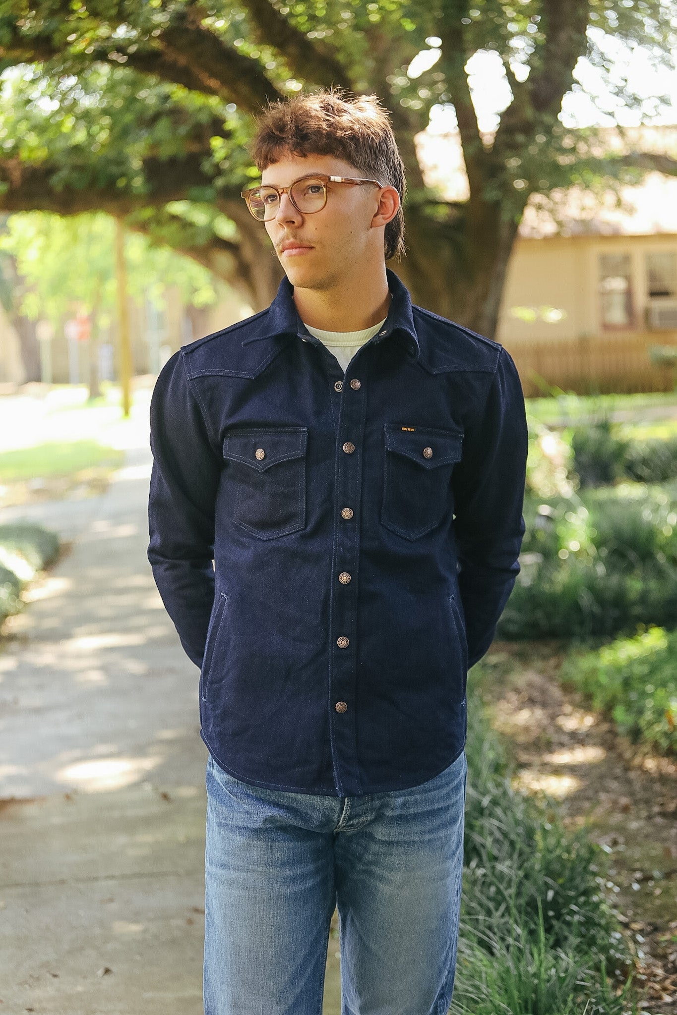A man wearing a dark navy blue button-up shirt jacket with two chest pockets and light blue jeans, standing outdoors on a paved path with trees and greenery in the background.