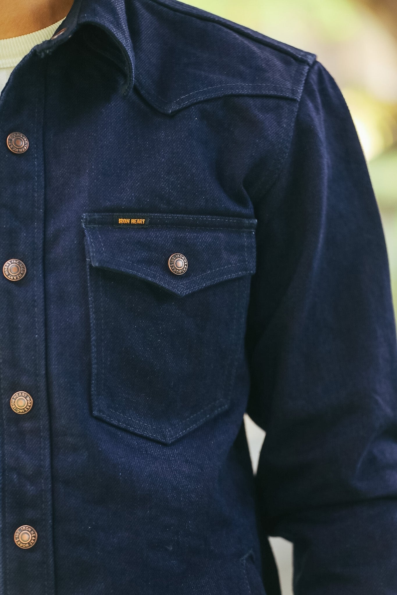 Close-up of a dark navy blue denim shirt jacket with a chest pocket and brass buttons, worn over a white shirt.