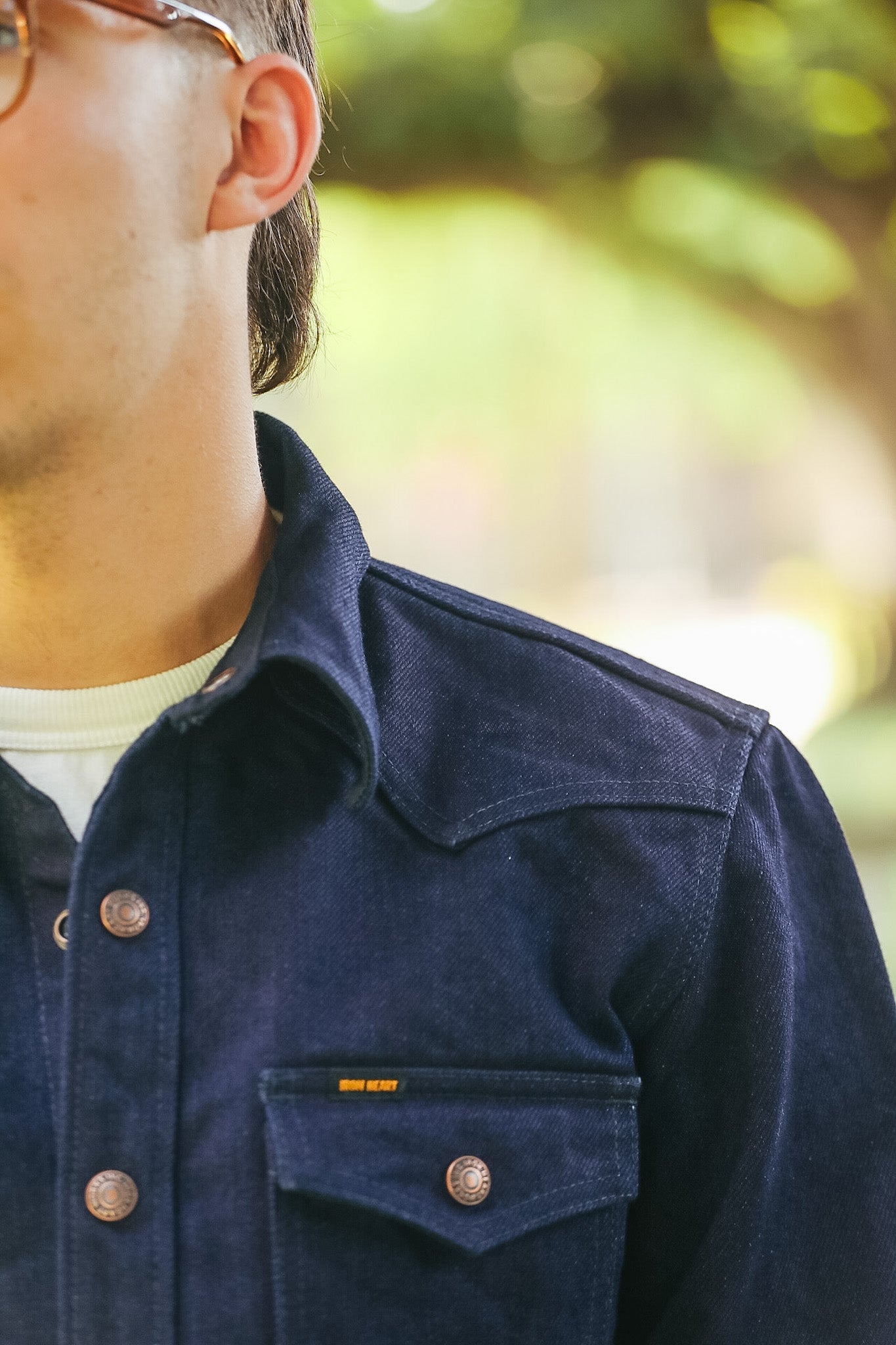 Close-up of a man wearing a dark navy blue denim button-up shirt with a chest pocket and a small orange 'BONSAI' label, set against a blurred green outdoor background.