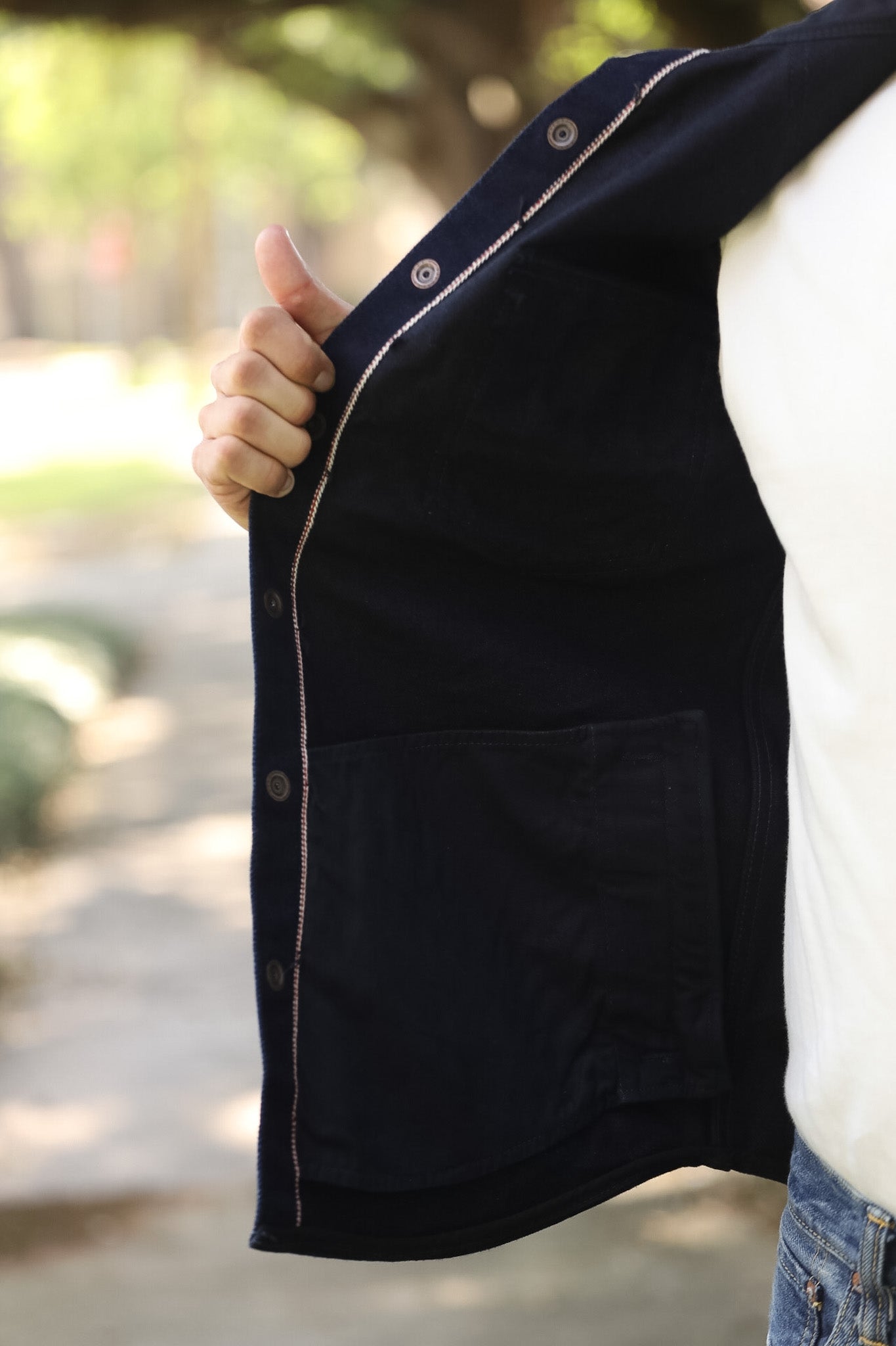 A close-up of a person outdoors holding open a dark navy jacket to show the interior lining and buttoned front edge. The person is wearing a white shirt and blue jeans, with a blurred park background.