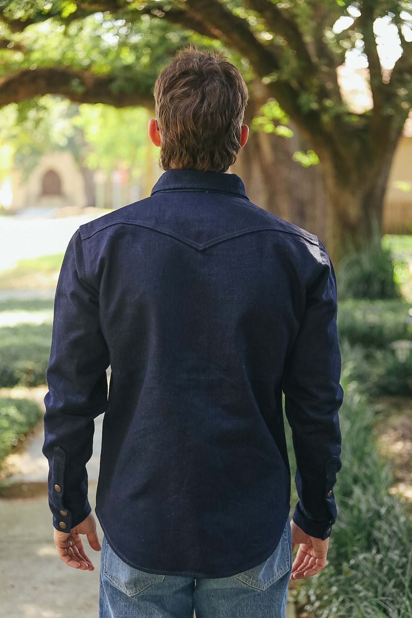 A man viewed from behind wearing a dark navy blue long-sleeve western-style shirt and blue jeans, standing outdoors in a park-like setting with trees and greenery in the background.