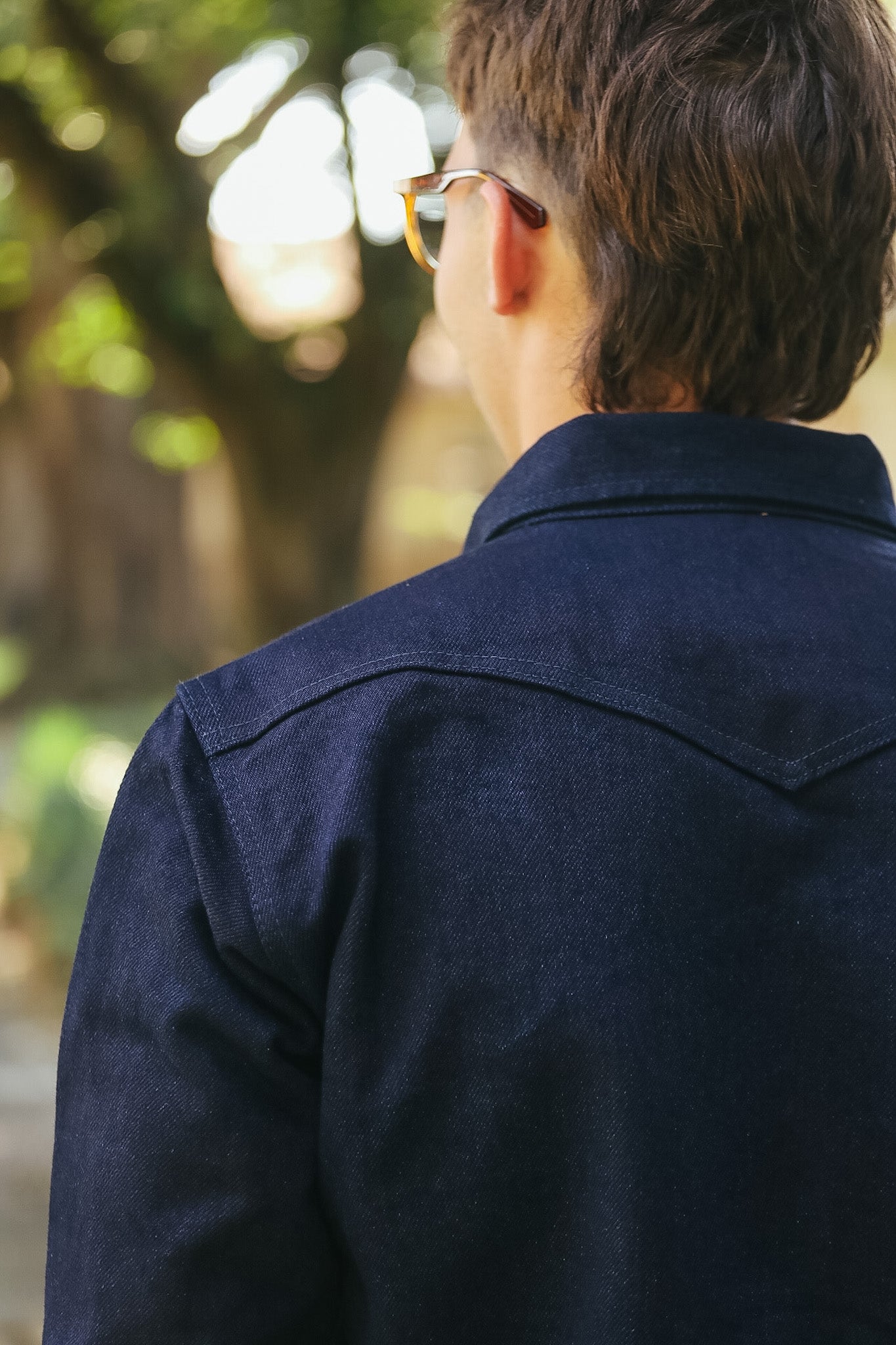 A man viewed from behind wearing a dark navy blue denim-style collared shirt and gold-framed sunglasses, standing outdoors with a blurred tree background.