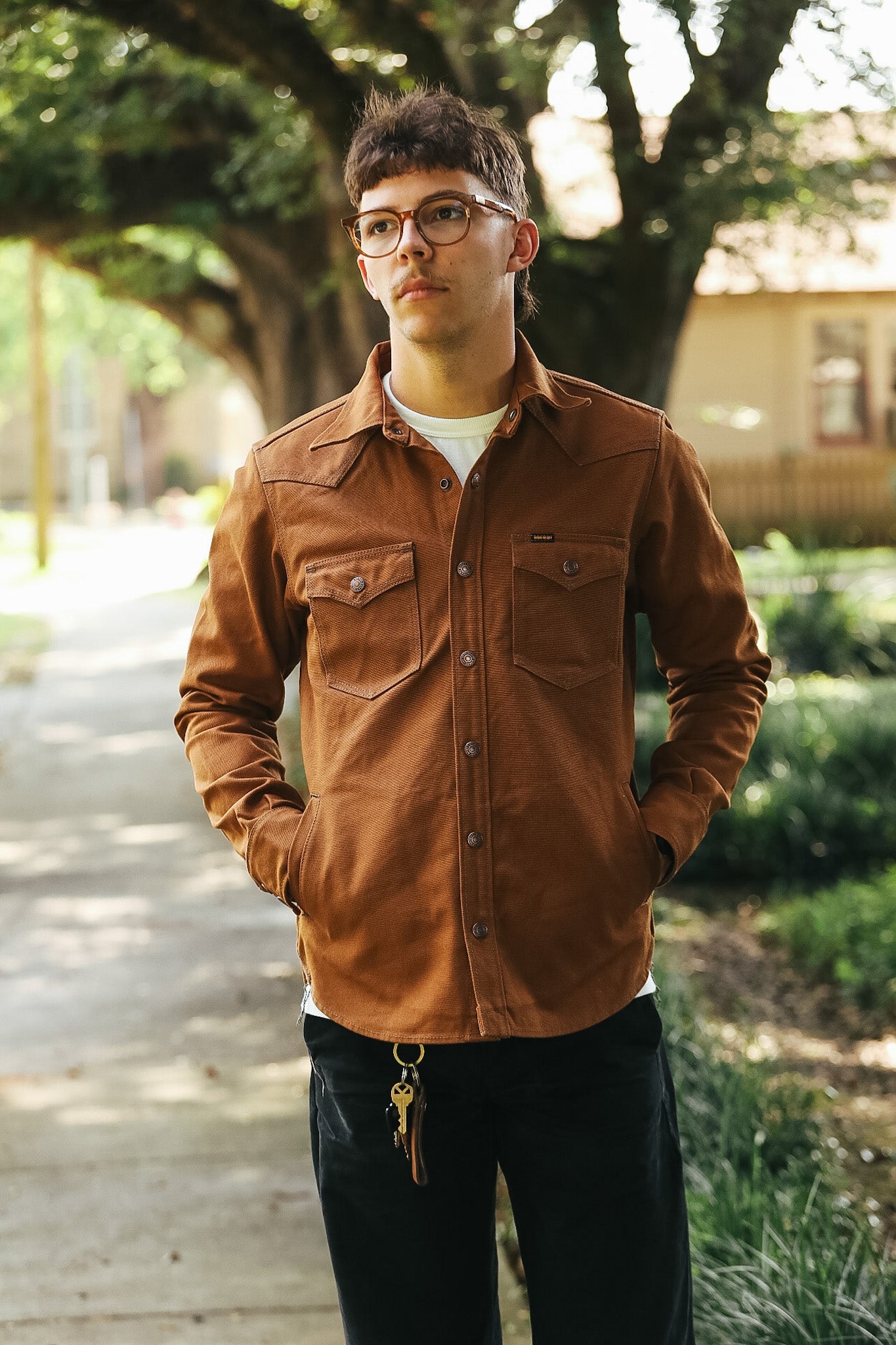 A man wearing a brown button-up shirt jacket over a white t-shirt and black pants, standing on a tree-lined path.
