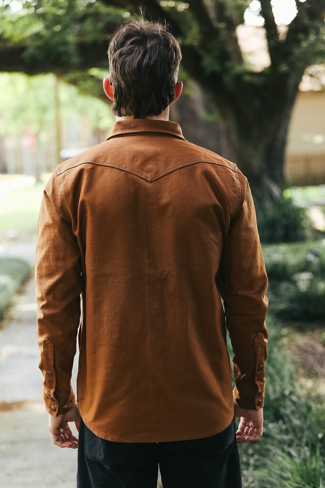 A man viewed from behind wearing a brown corduroy button-down shirt and dark pants, standing outdoors on a path with trees and greenery in the background.