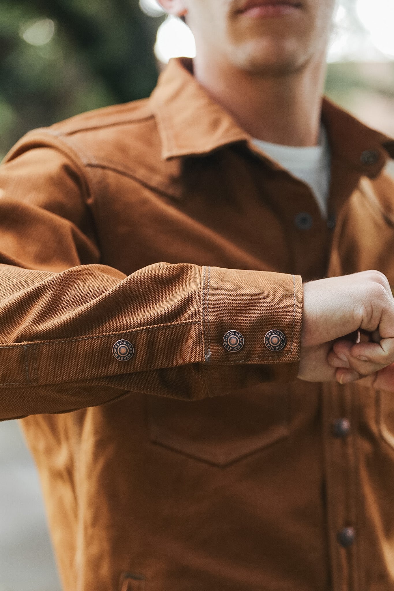 A close-up of a person wearing a brown suede or nubuck utility jacket over a white t-shirt, with the person's hands adjusting the cuff of the jacket.