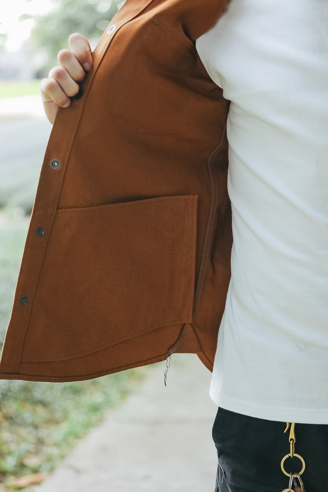 A close-up lifestyle shot of a person wearing a brown canvas utility jacket over a white t-shirt, holding the jacket open to show the interior pocket and snap-button closure.