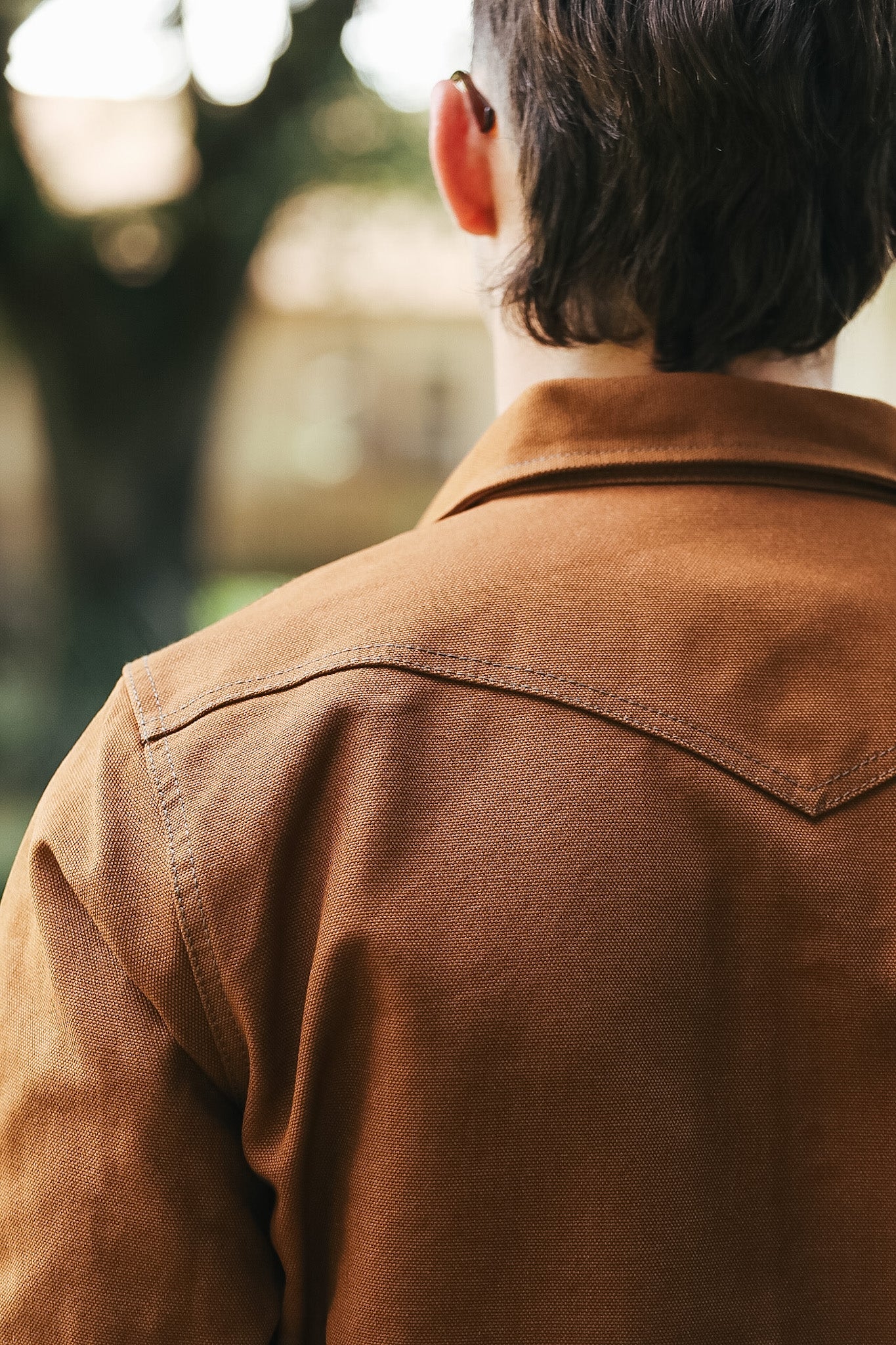 A close-up back view of a person wearing a brown canvas utility jacket, with a softly blurred outdoor background.