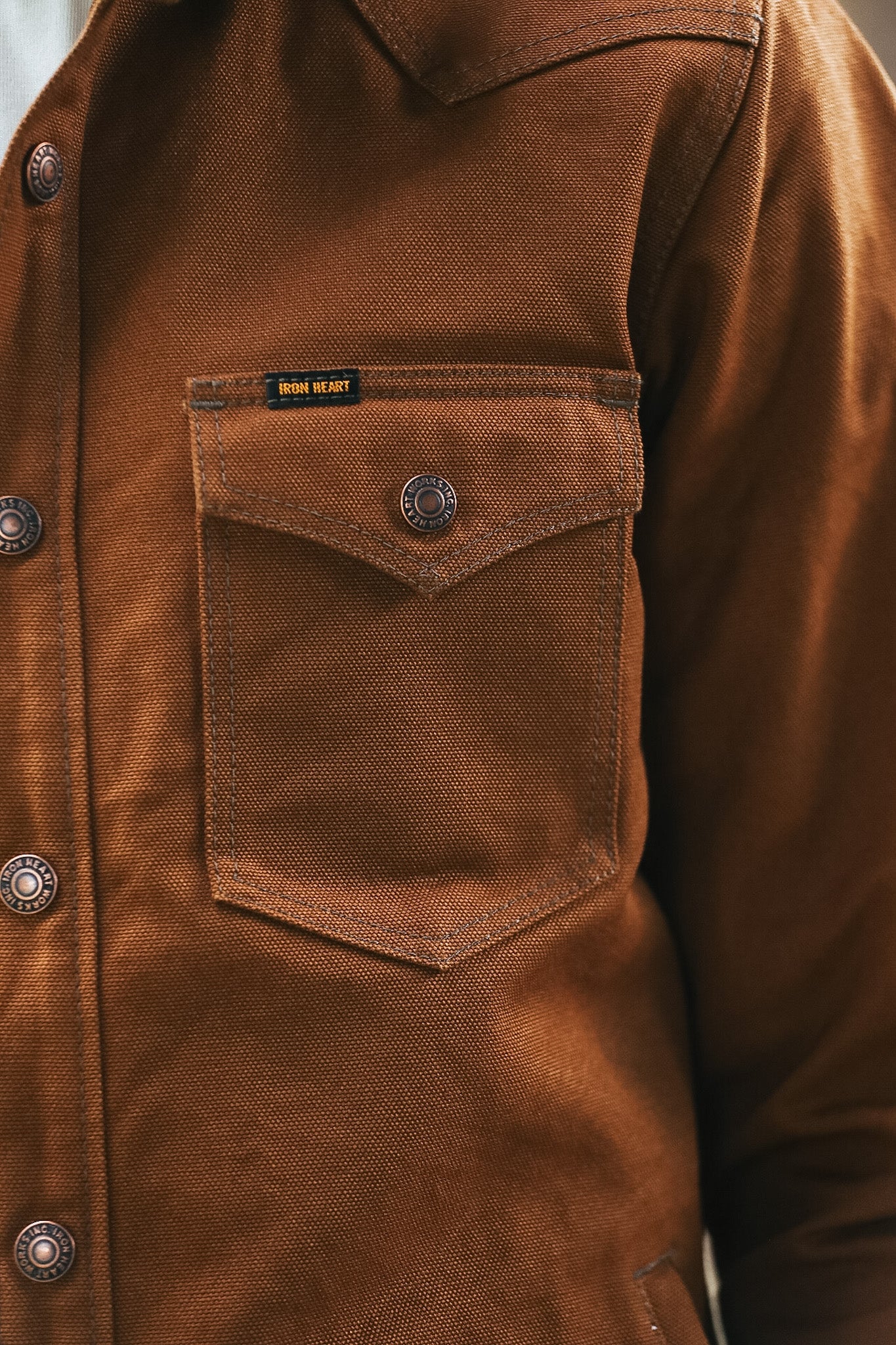 Close-up of a brown canvas work jacket with a chest pocket and metal snap buttons, showing a small black and yellow 'BORN HEART' label.