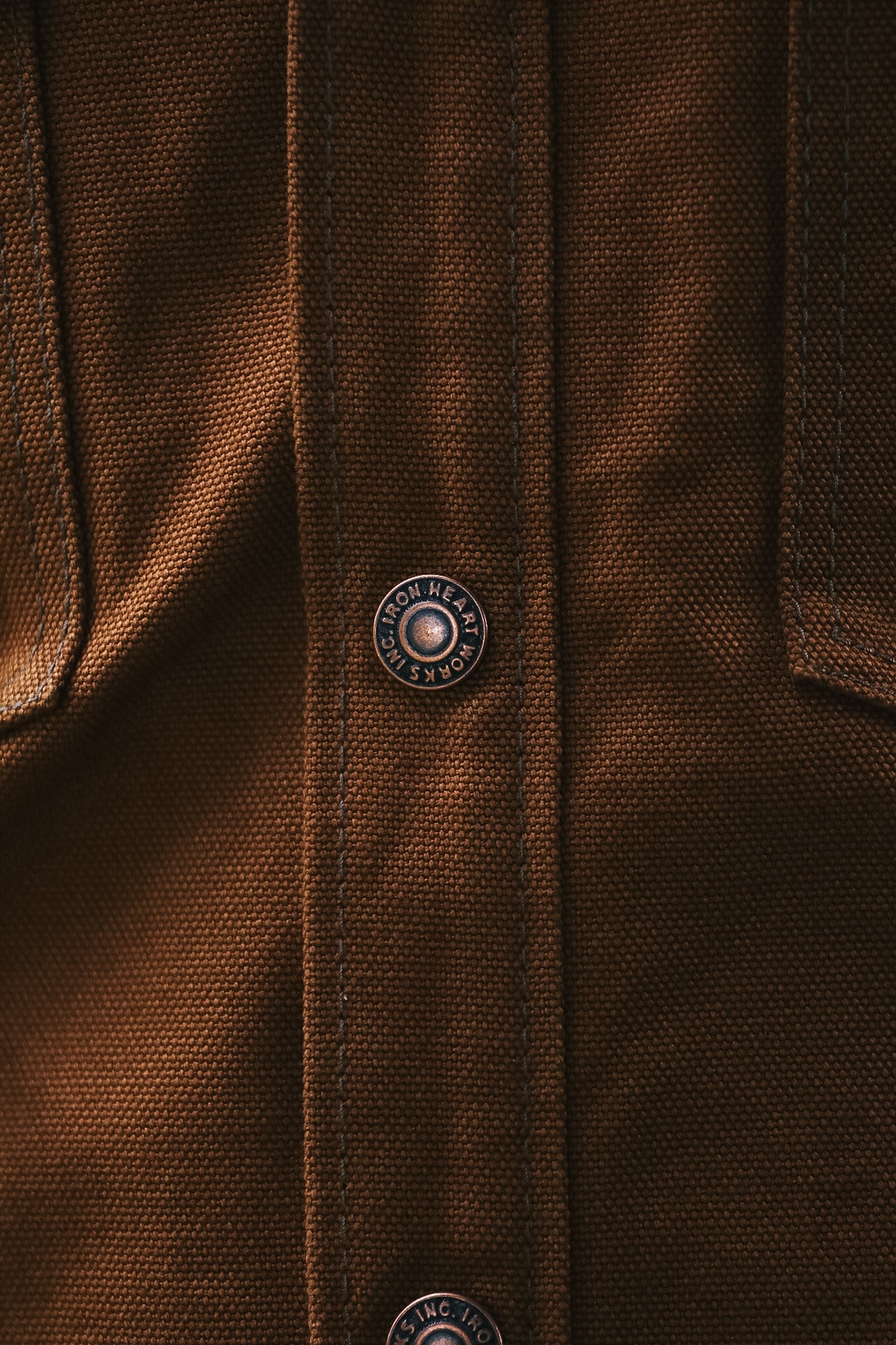 Close-up of a brown textured fabric garment with a vertical button placket and a visible round metal button engraved with the brand name 'HARROSH'.
