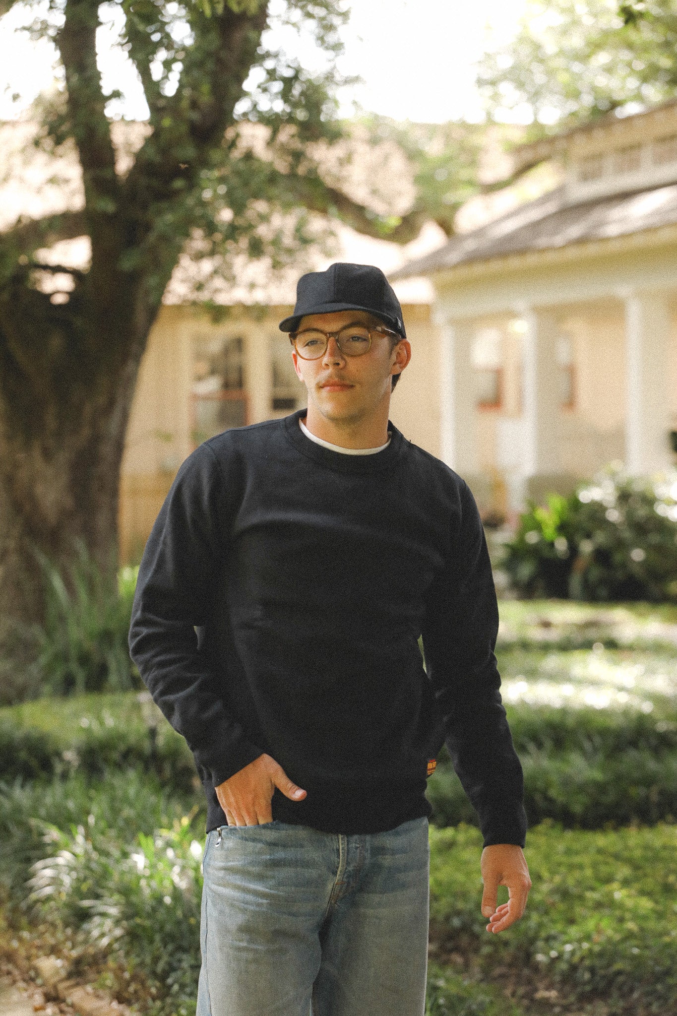 Man wearing a black sweater and cap standing outdoors with a house and trees in the background