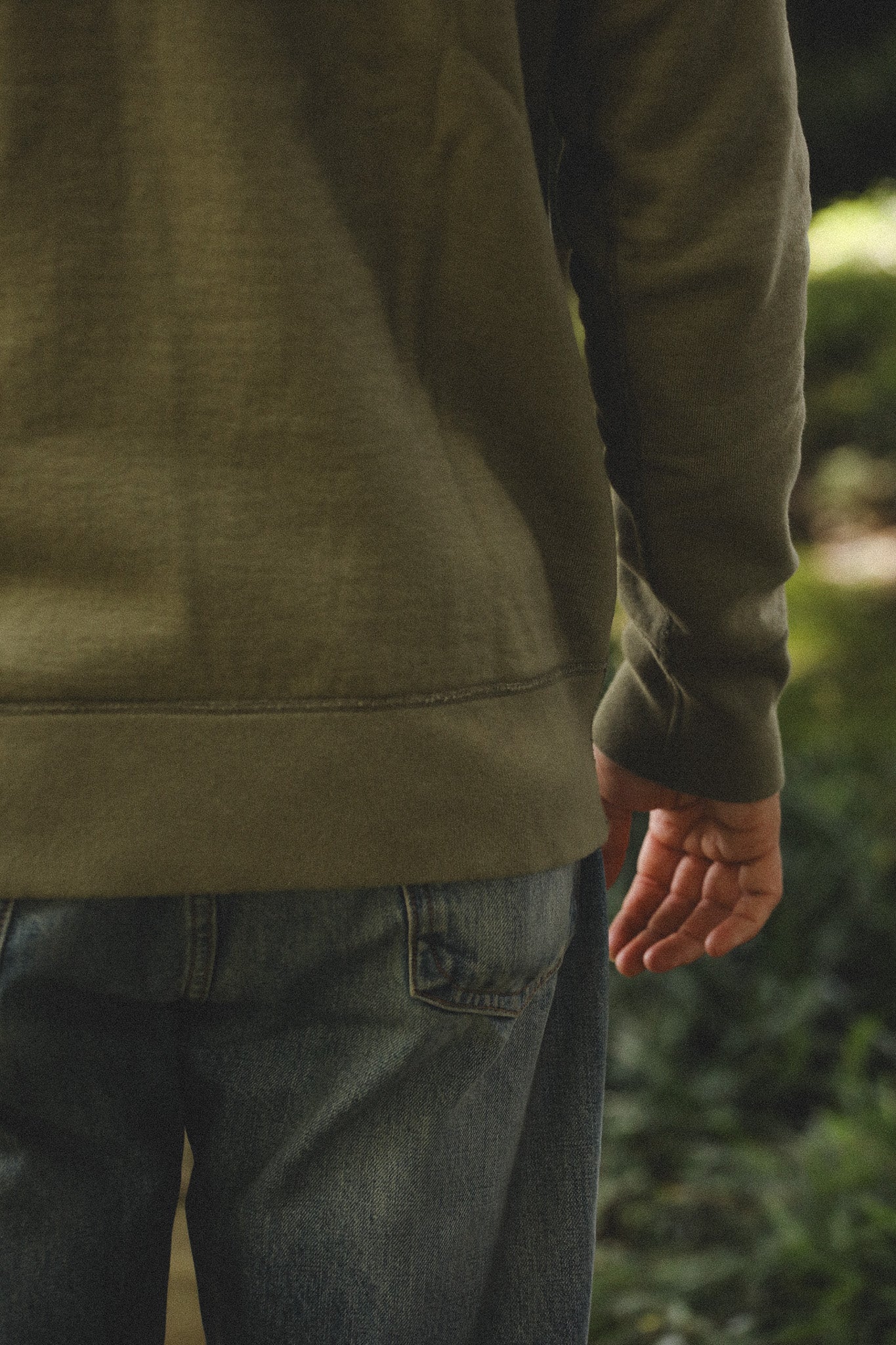 Person wearing a green sweatshirt and jeans standing outdoors with a blurred natural background