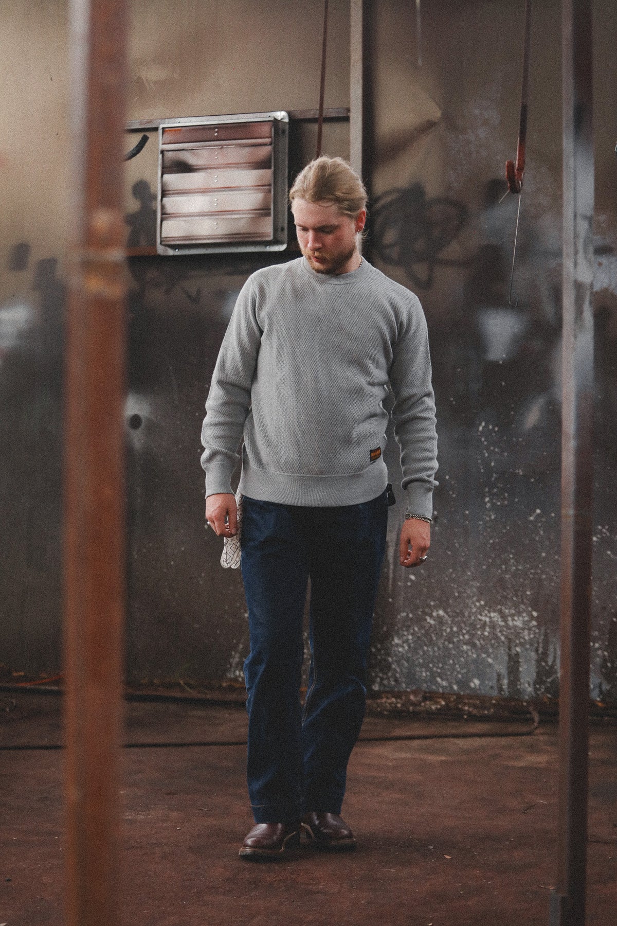 A man wearing a light gray crewneck sweatshirt, dark blue jeans, and brown shoes stands in a gritty, industrial room with graffiti on the walls.