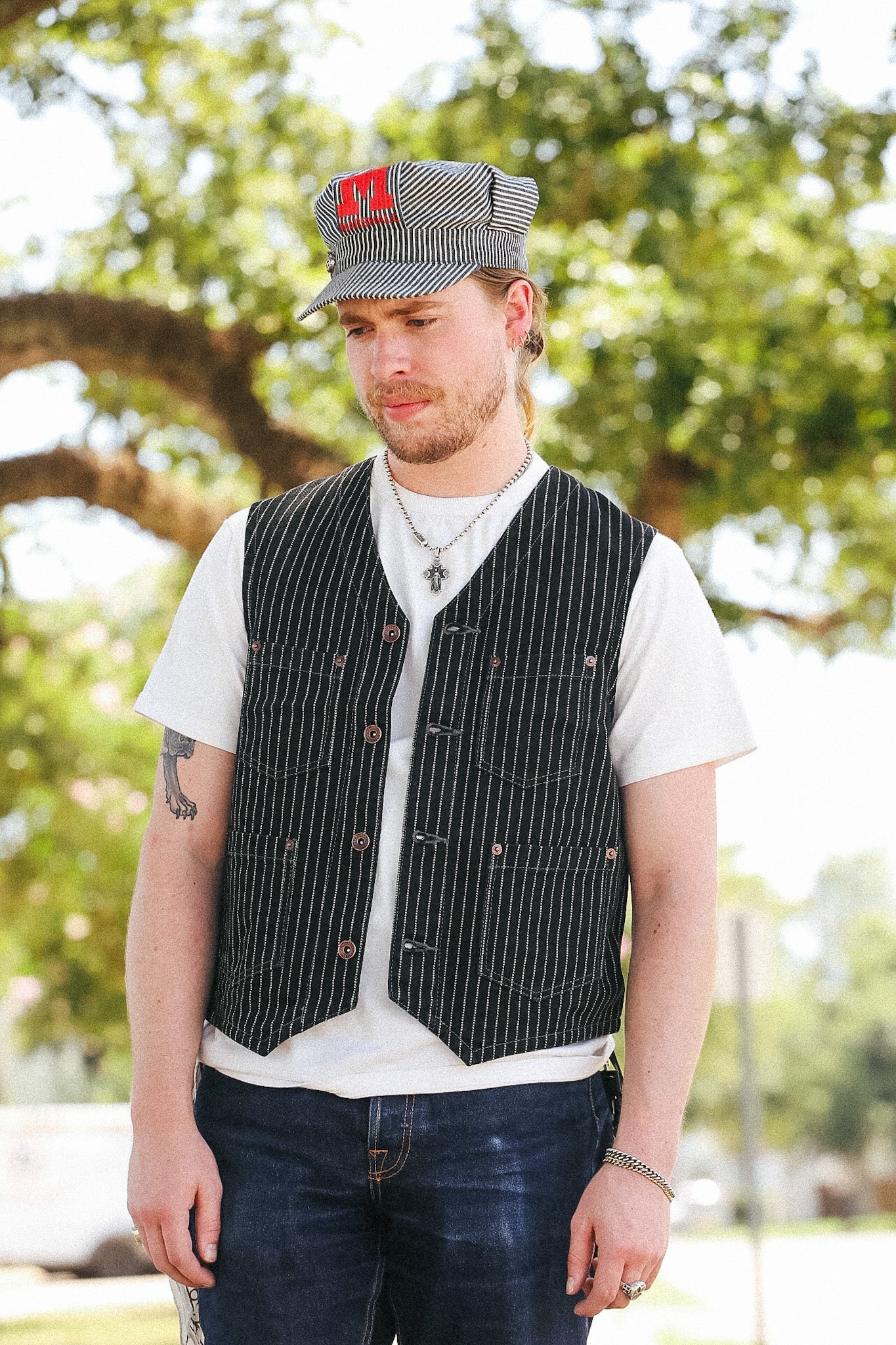 A man wearing a black and white pinstriped vest over a white t-shirt, dark jeans, and a gray and red striped railroad conductor hat, standing outdoors in front of a blurred green tree.