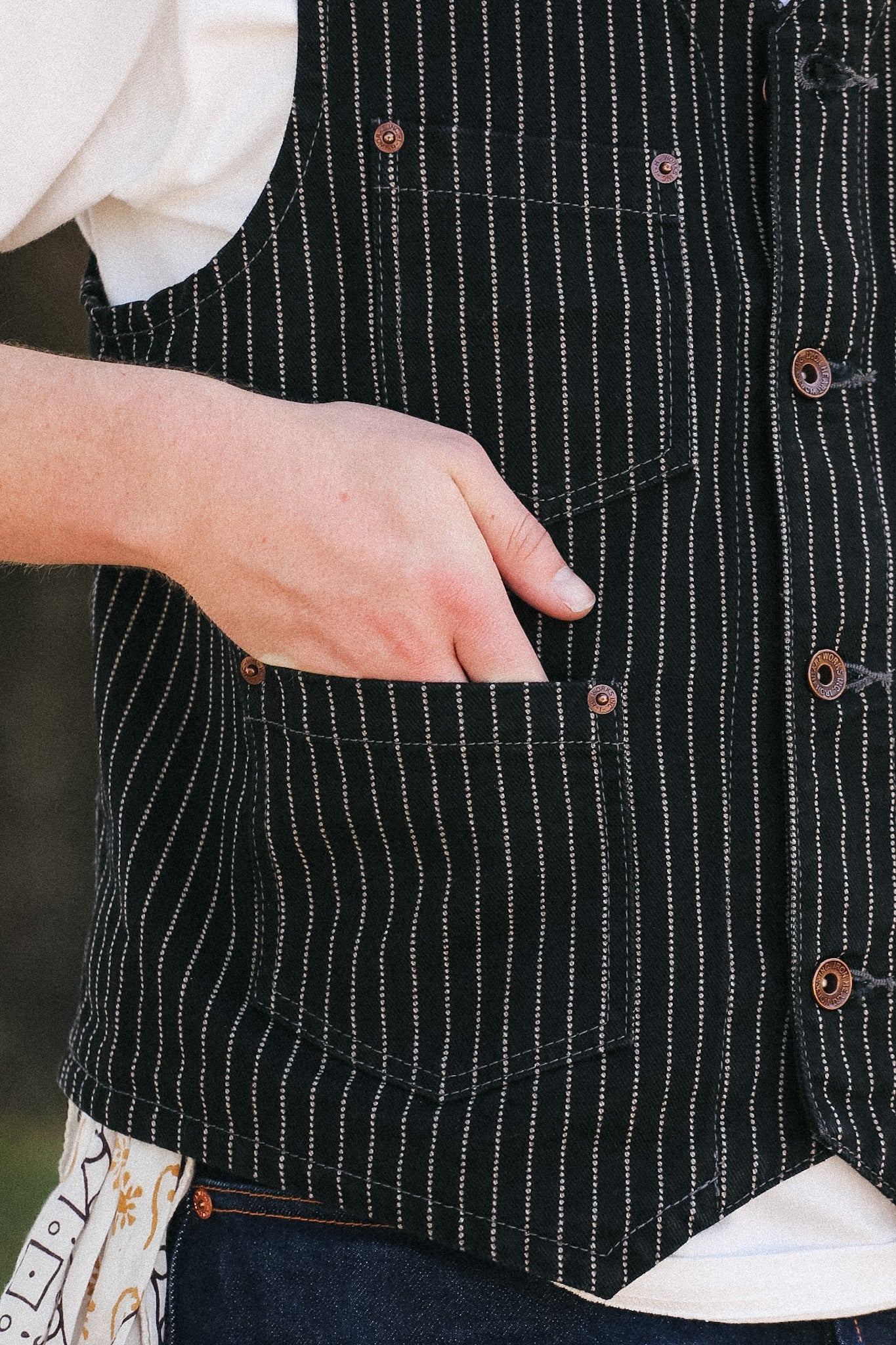 Close-up of a person wearing a black vest with thin white vertical pinstripes, brown buttons, and a front patch pocket, with a hand reaching into the pocket.