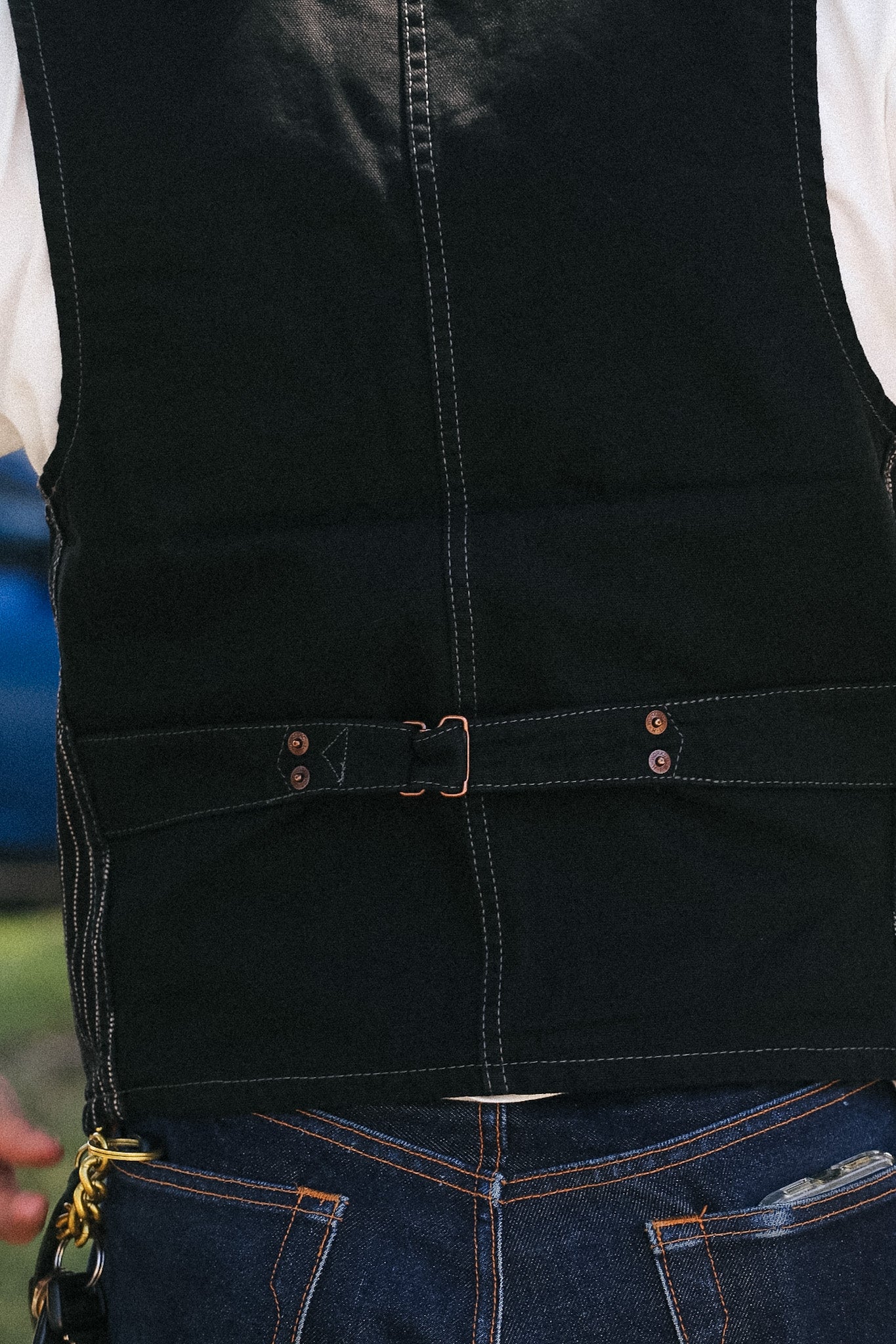 Close-up of a black denim vest with a matching belt and buckle, worn over a white shirt and blue jeans.
