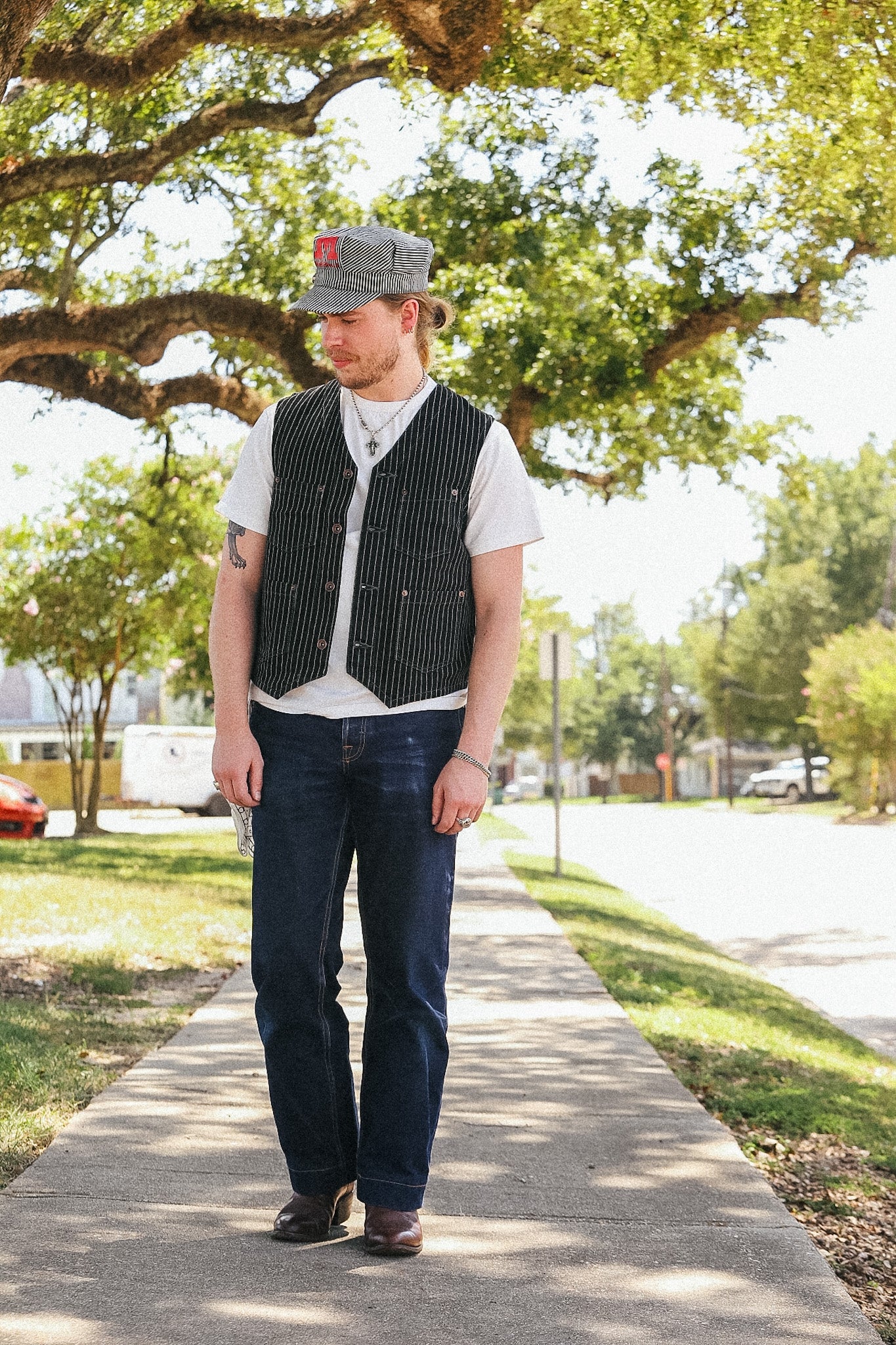 A man standing on a tree-lined sidewalk wearing a gray pinstripe vest over a white t-shirt, dark blue jeans, and a gray baseball cap.