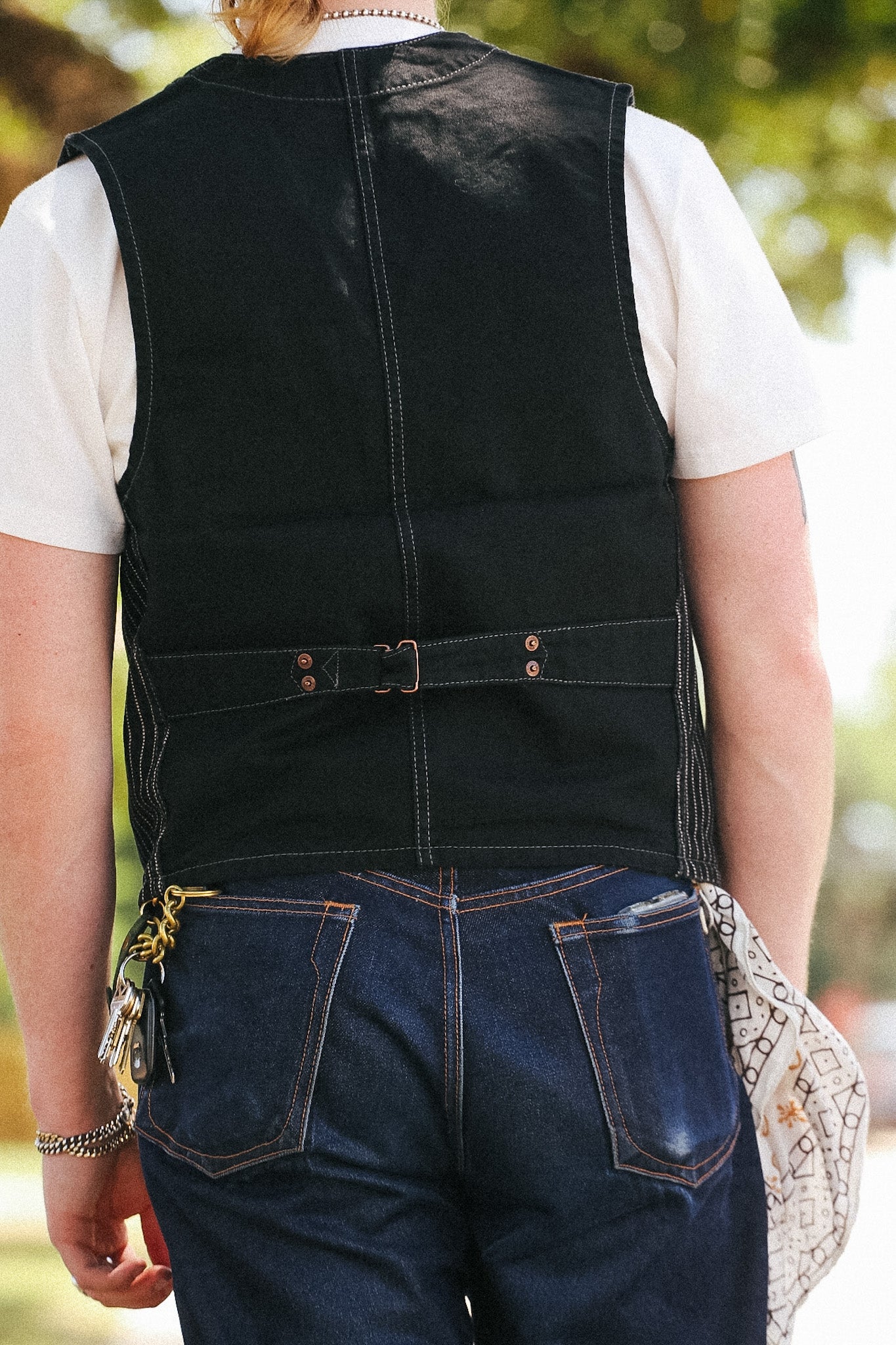 A person shown from behind wearing a black denim vest over a white t-shirt and dark blue jeans, with a patterned clutch bag hanging from their hand, set against a blurred outdoor background.