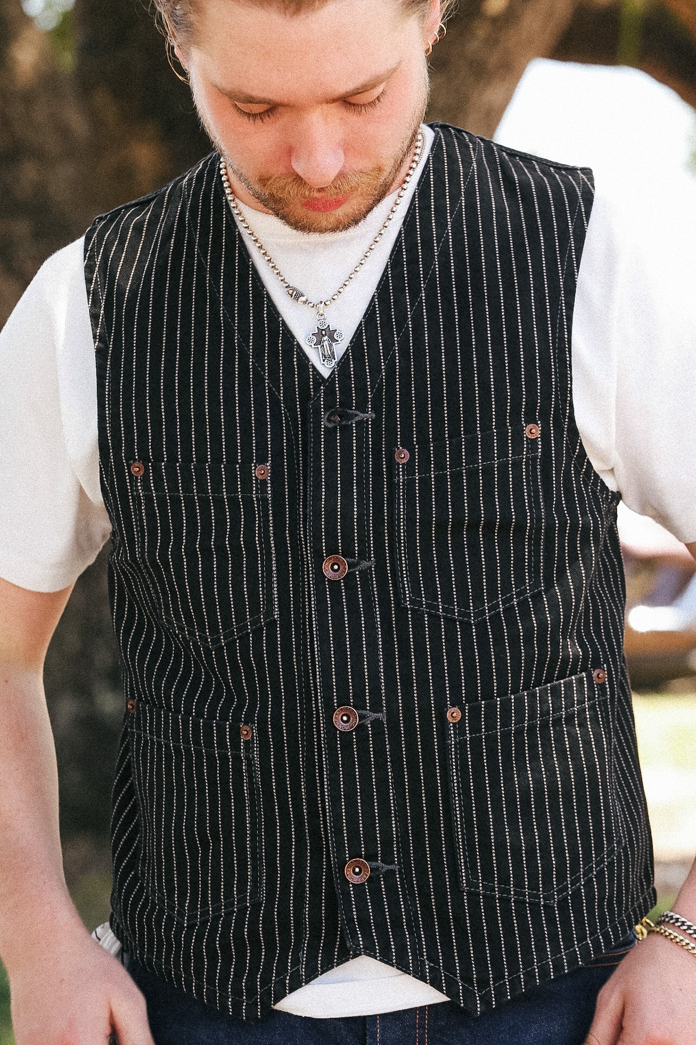 A man wearing a black pinstriped vest over a white t-shirt and blue jeans, looking down. The vest features a button front and four front pockets.