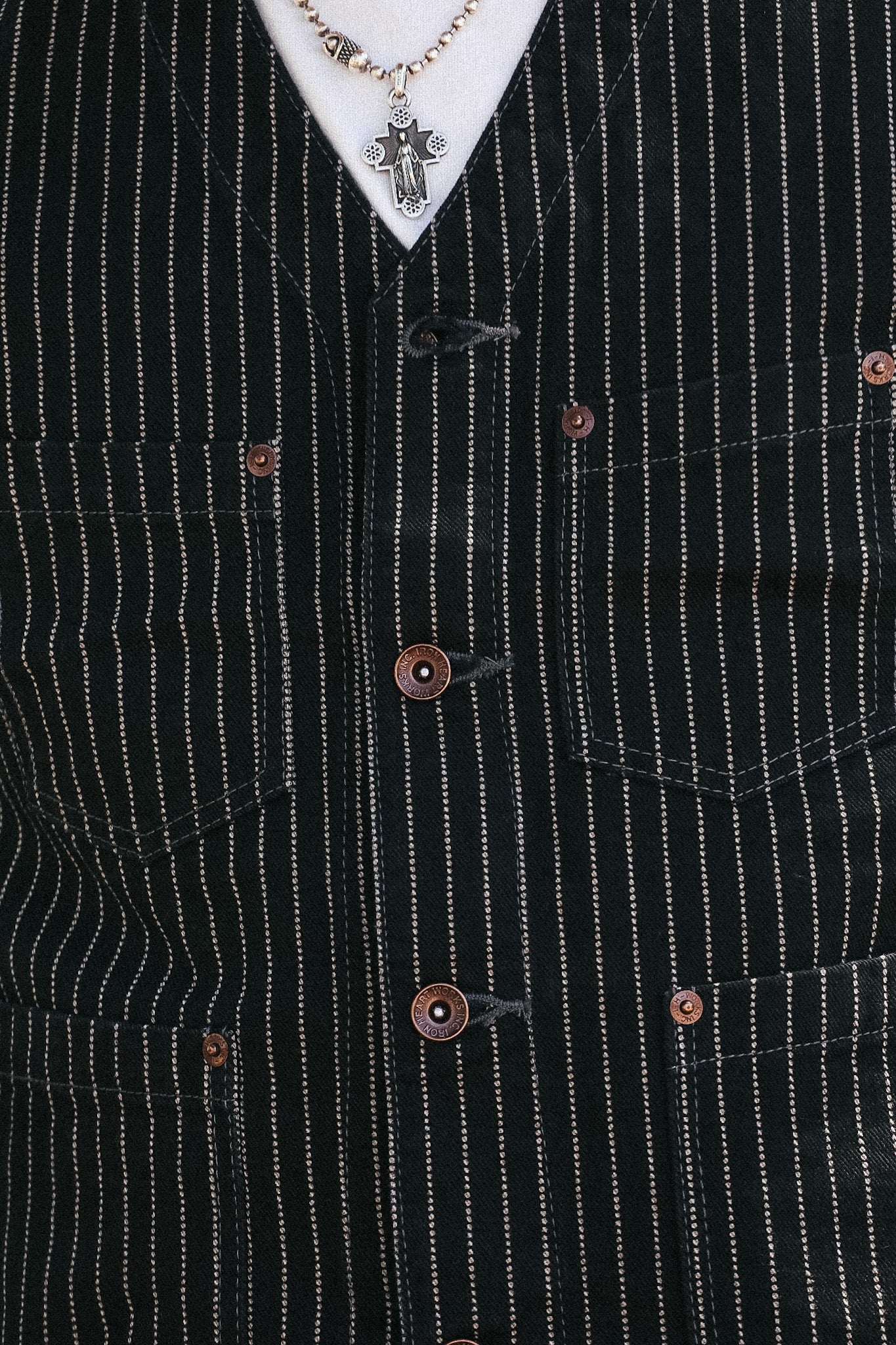 Close-up of a black pinstripe vest with copper-colored buttons, worn over a white shirt and a silver cross pendant necklace.