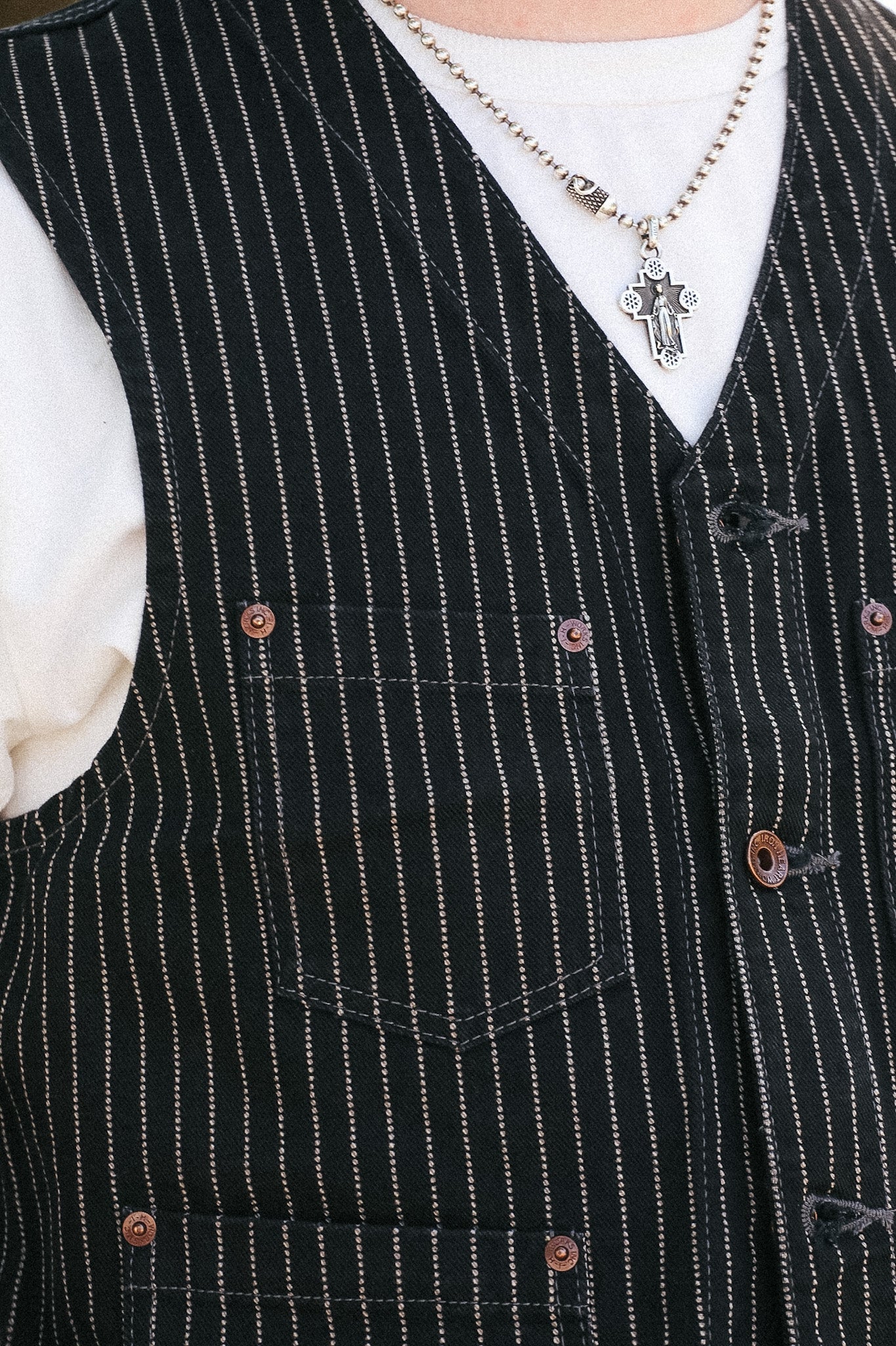 Close-up of a black pinstripe vest with brown buttons, worn over a white t-shirt and a silver cross necklace.