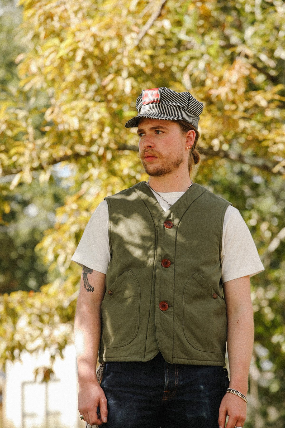 A man wearing an olive green button-front vest over a white t-shirt and a gray-and-white striped cap, standing outdoors in front of a blurred background of green and yellow autumn trees.