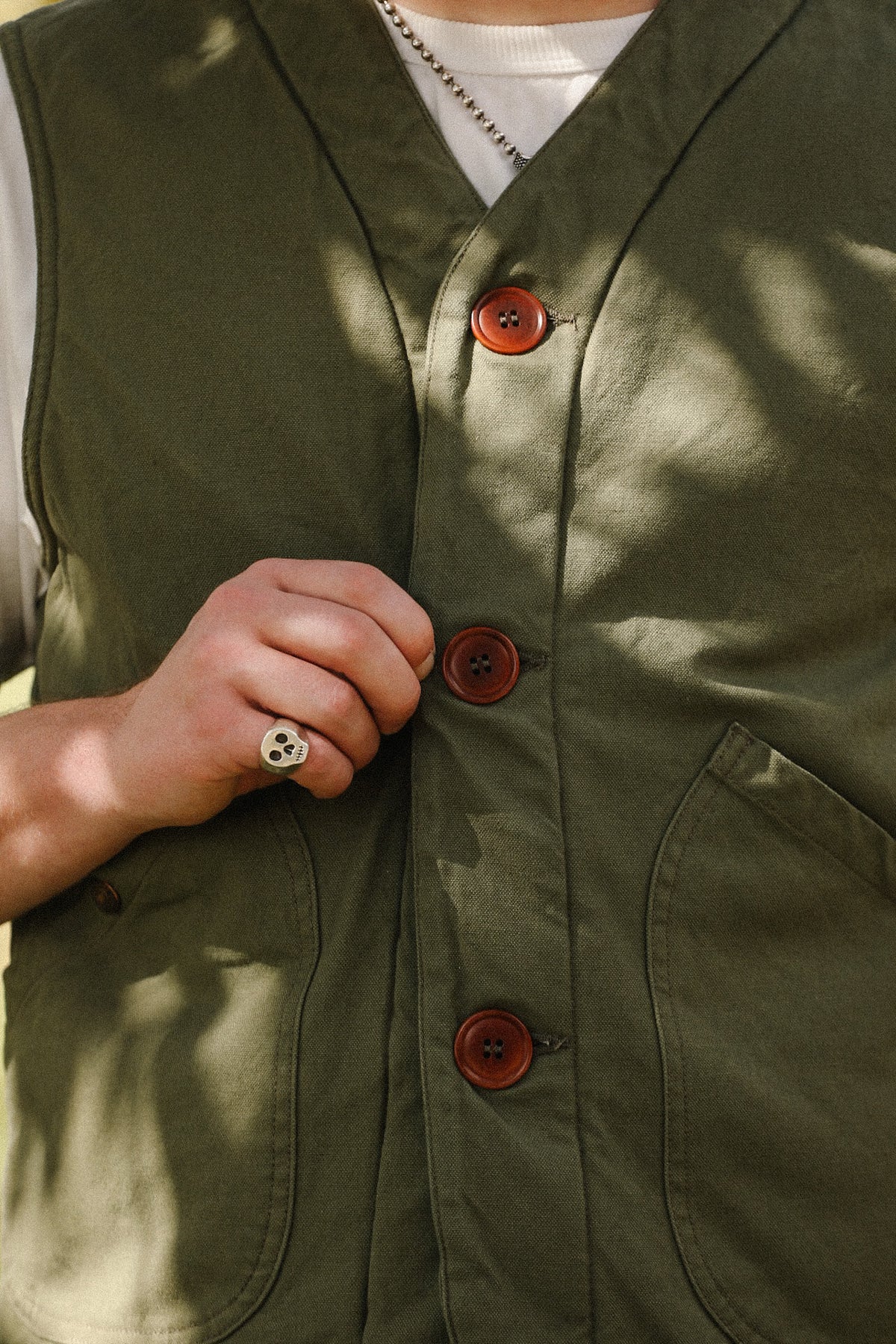 Close-up of a person wearing an olive green button-front vest over a white shirt, with a hand adjusting one of the brown buttons. The photo is lit by dappled sunlight and shadows.